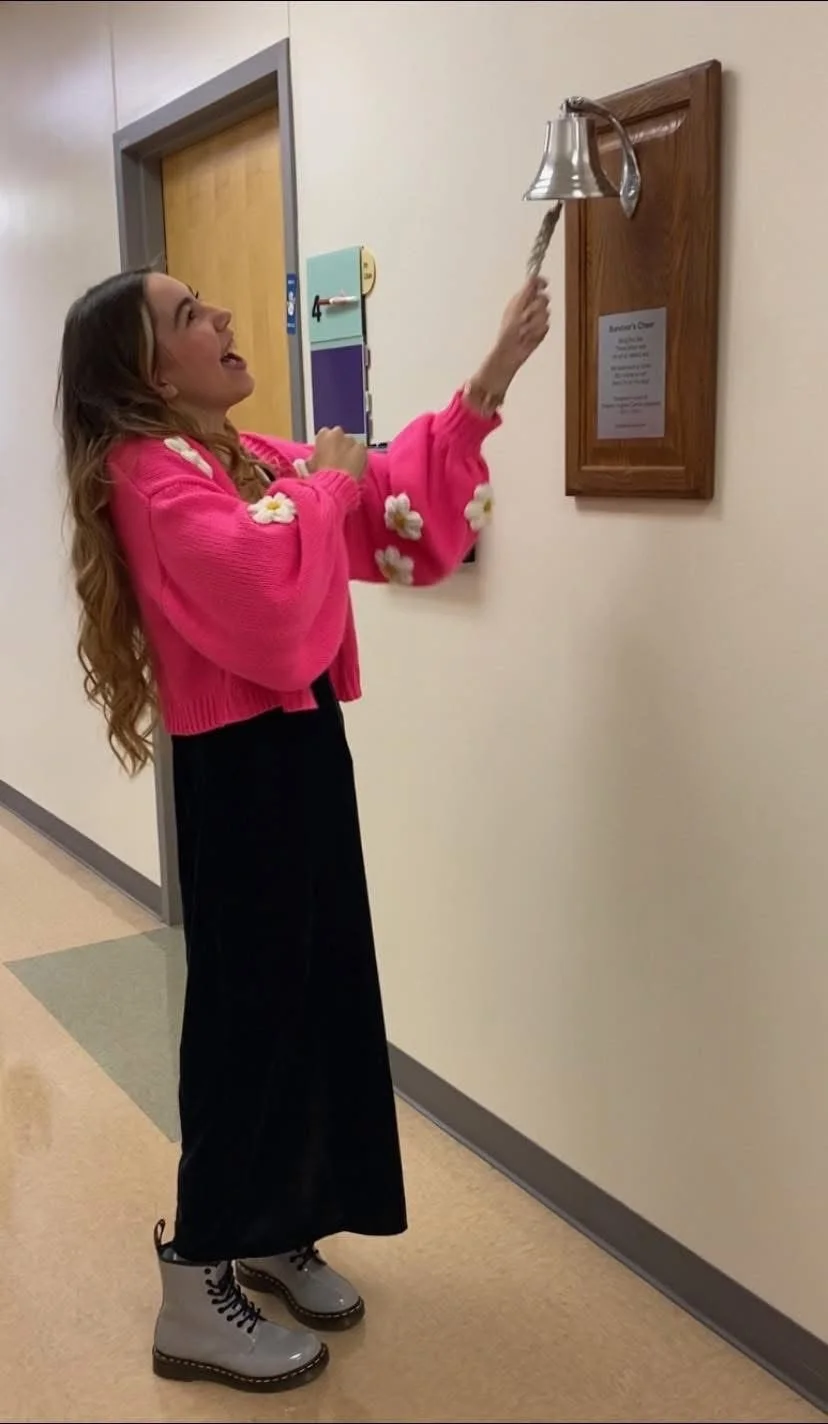 A woman with long hair wearing a pink sweater with flower decorations, black long skirt, and gray boots, playfully ringing a bell mounted on a wooden plaque with a rope handle on a beige wall.