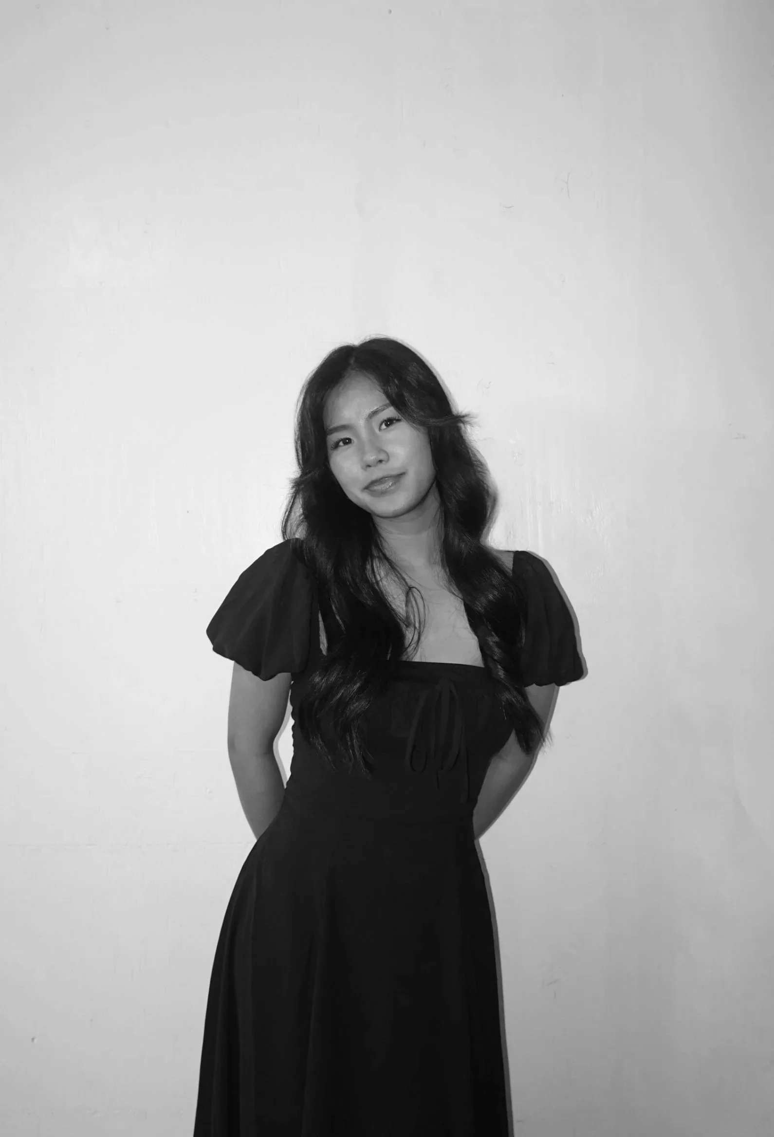 A woman with long wavy hair in a black dress with puffed sleeves, standing against a plain wall, smiling slightly.