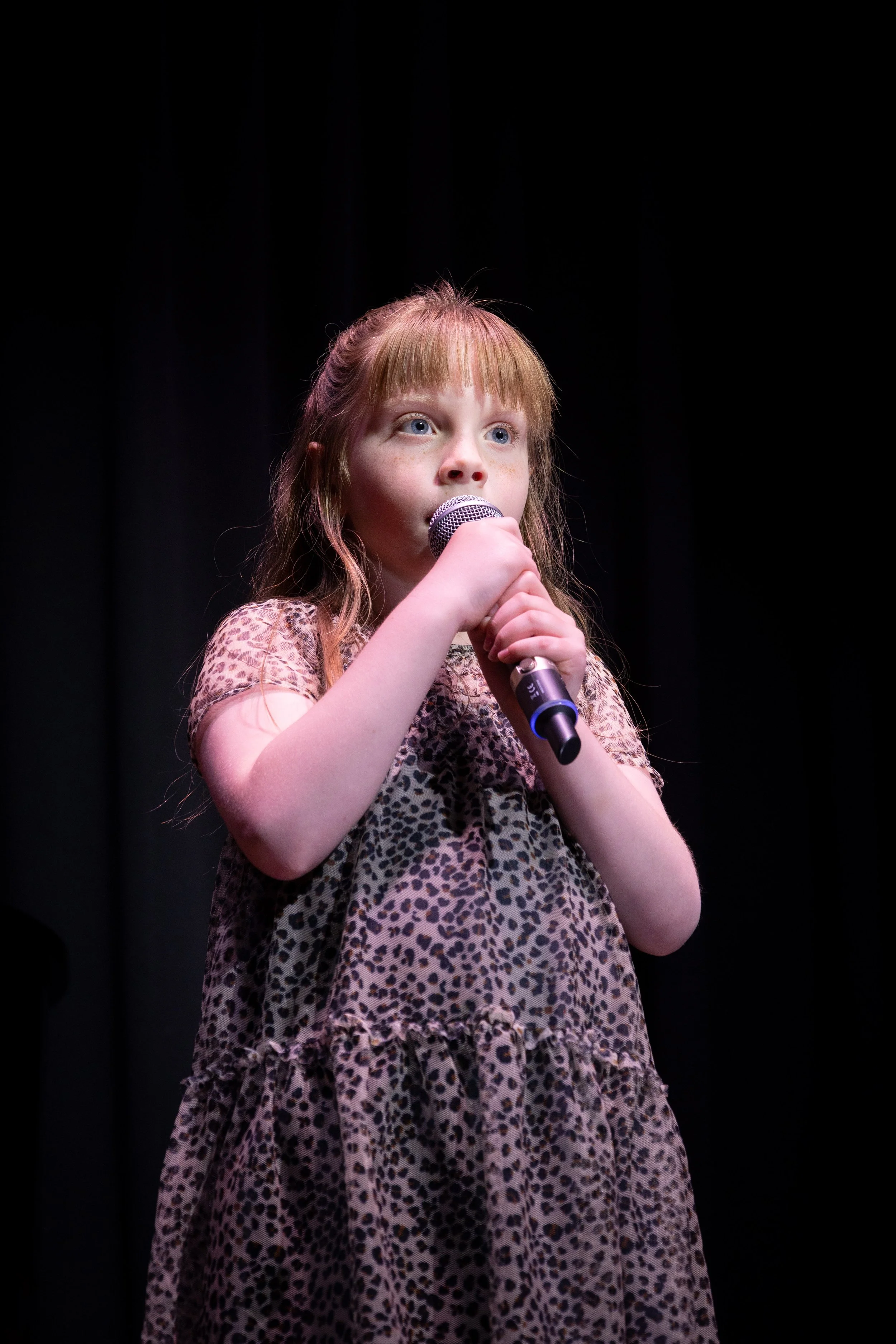 A young girl with red hair and wearing a leopard print dress holding a microphone on stage in front of a black background.
