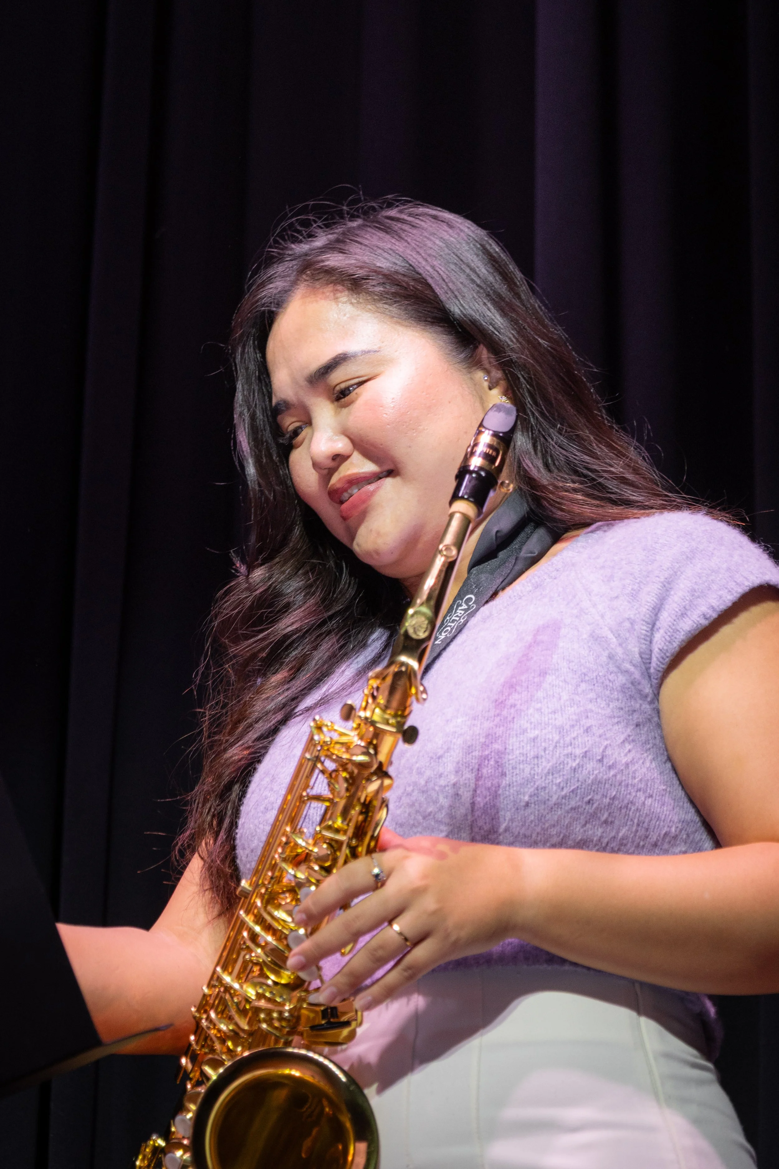 A woman with long dark hair playing a gold saxophone, smiling with her eyes closed, wearing a lavender top and a black scarf against a dark background.