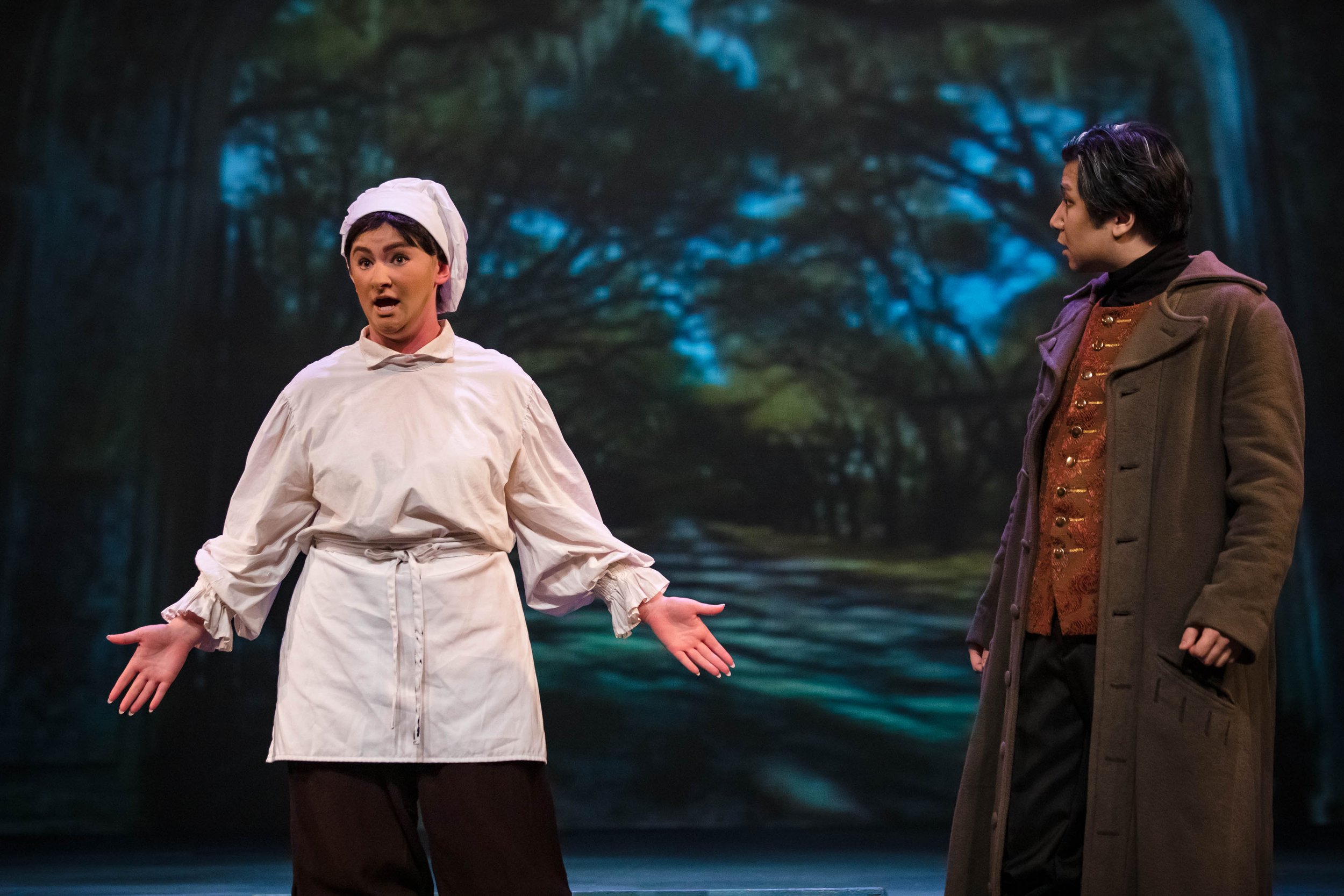 Two actors on stage during a theater performance; one dressed as a chef with a white coat and hat, the other in a long brown coat with a patterned waistcoat. They are in front of a backdrop of a forest scene.