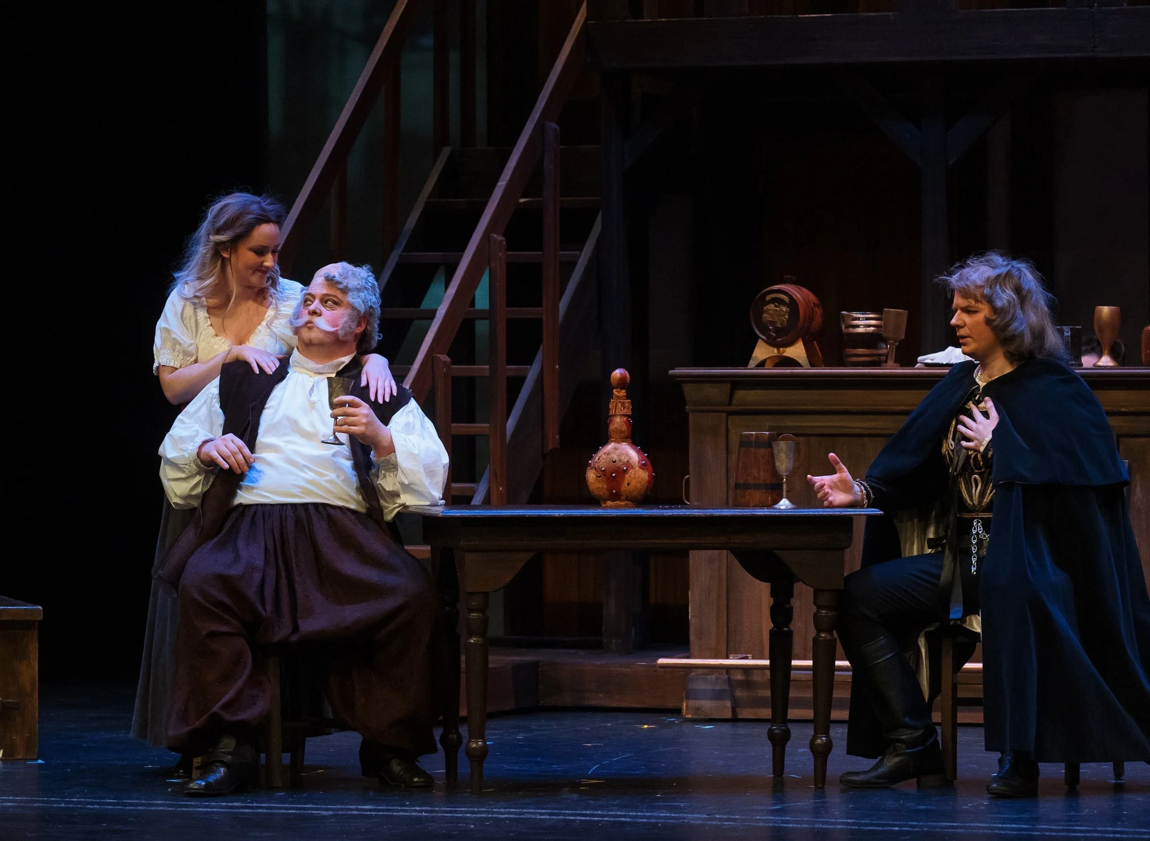 Scene from a theatrical play with three actors, one sitting and two standing, in period costumes, in a room with wooden furniture and staircase.