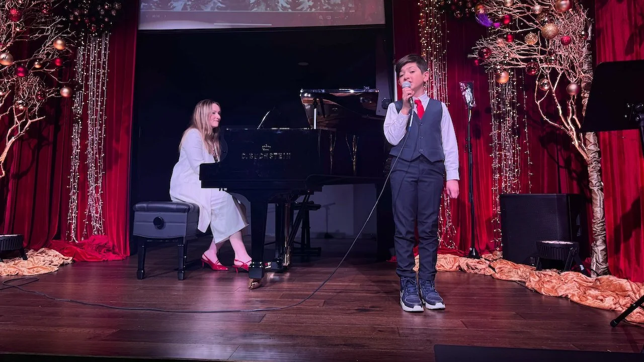 A young boy wearing a vest and red tie singing into a microphone on stage, with a woman playing the piano beside him. The stage is decorated with red curtains and gold and purple ornaments.