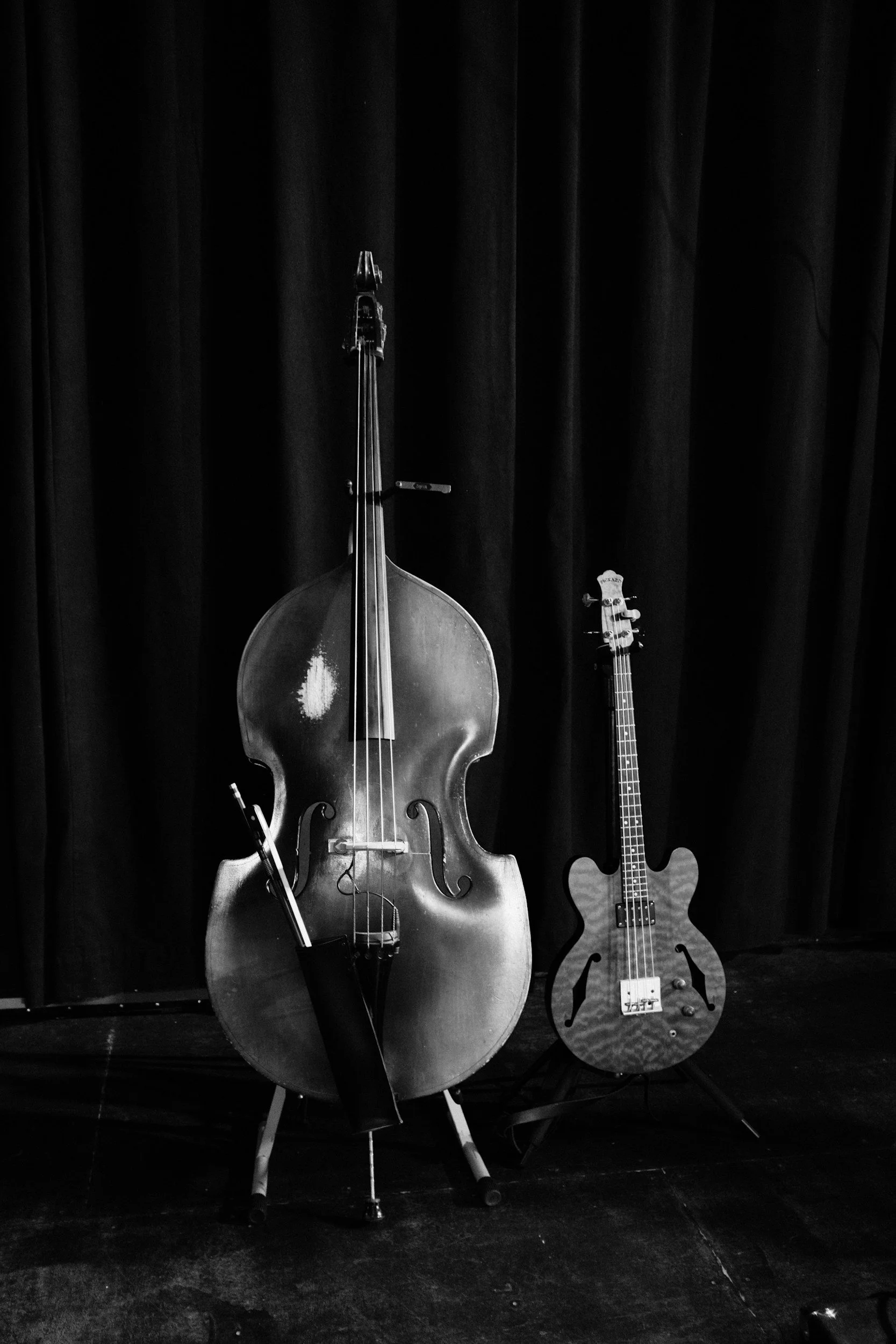 Black and white photo of a double bass and an electric guitar on a stage with a dark curtain background.