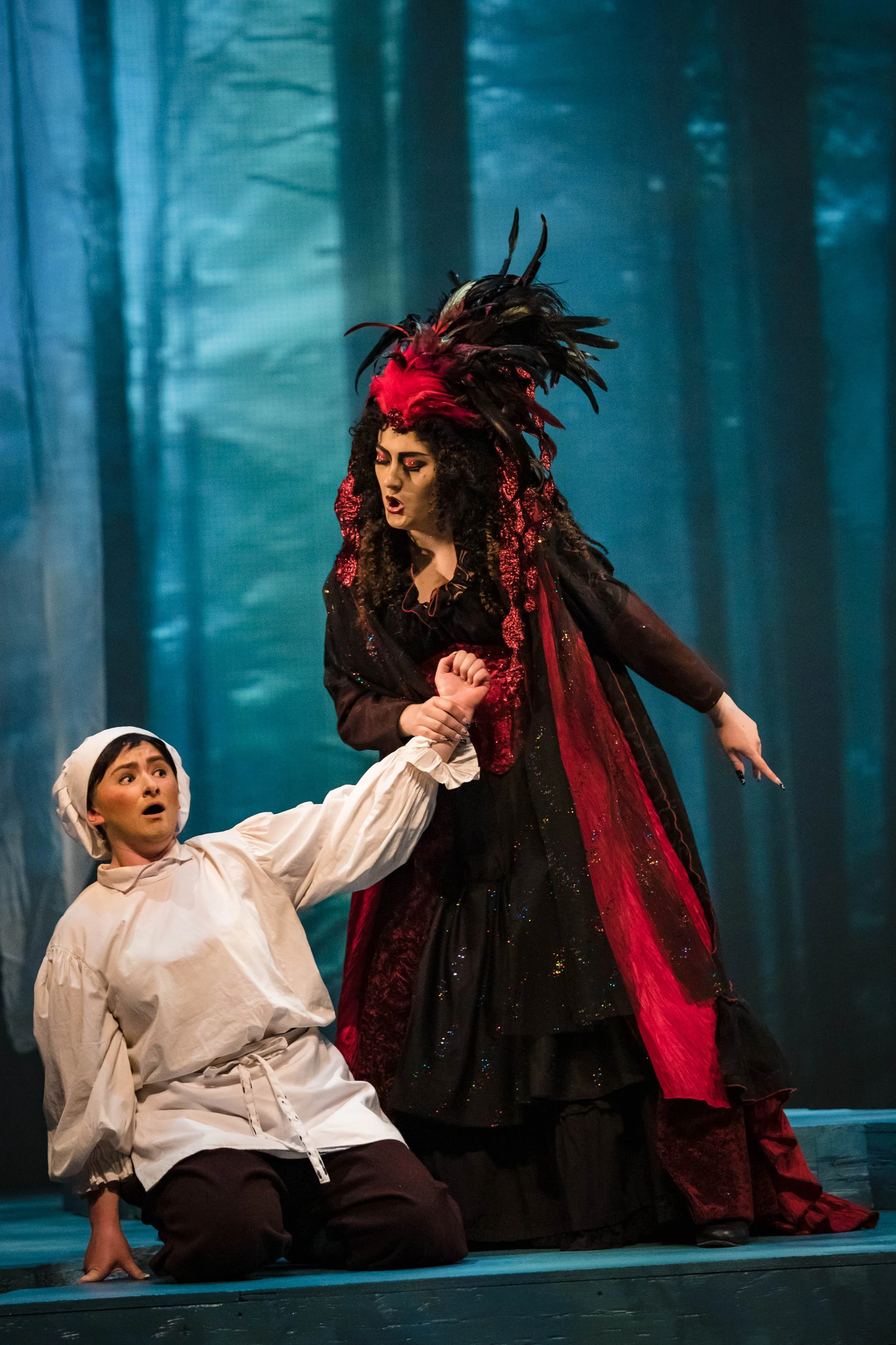 Two women dressed in costumes, one in a peasant outfit and the other in a flamboyant, dark costume with a feathered headdress, appear to be performing a theatrical scene in a forest setting.