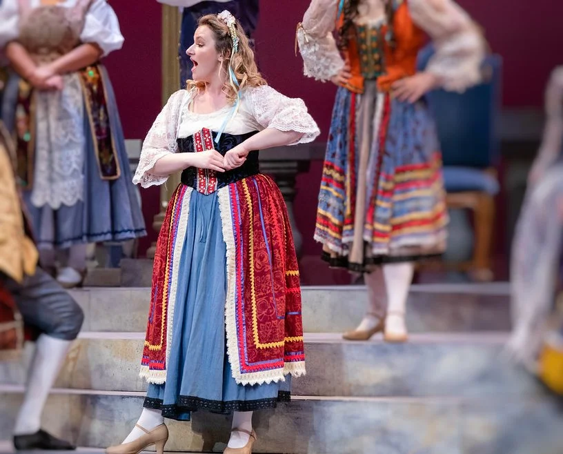 A woman in traditional folk costume singing or performing on stage, surrounded by similarly dressed people.