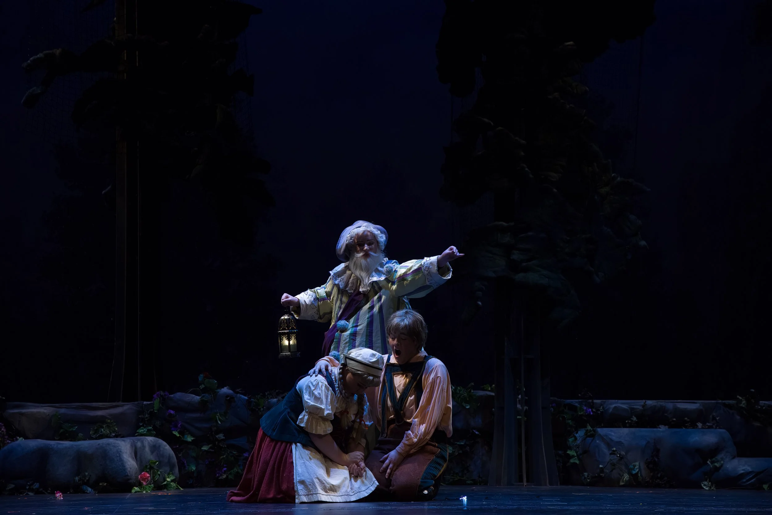 Three actors on stage, one dressed as an old man with a lantern, two children kneeling, in a forest scene with trees and rocks.