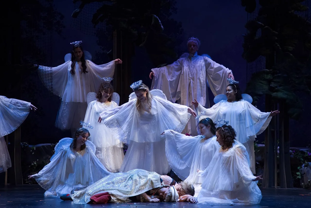 A stage scene from a Christmas play depicting angels in white costumes with halos, surrounding a girl laying on the ground, possibly portraying a nativity scene.