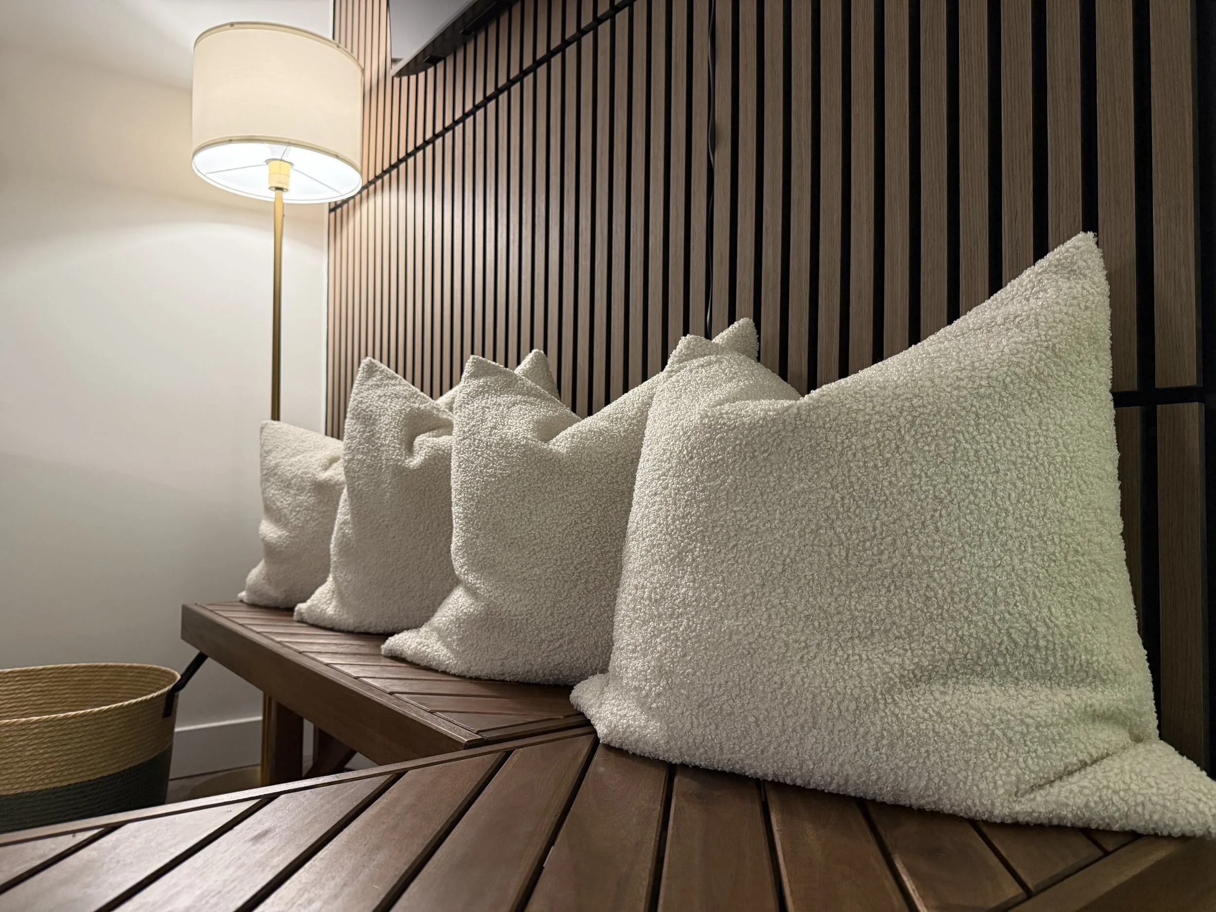Four white textured pillows on a wooden bench next to a wall with wood paneling, with a lamp and a woven basket nearby.