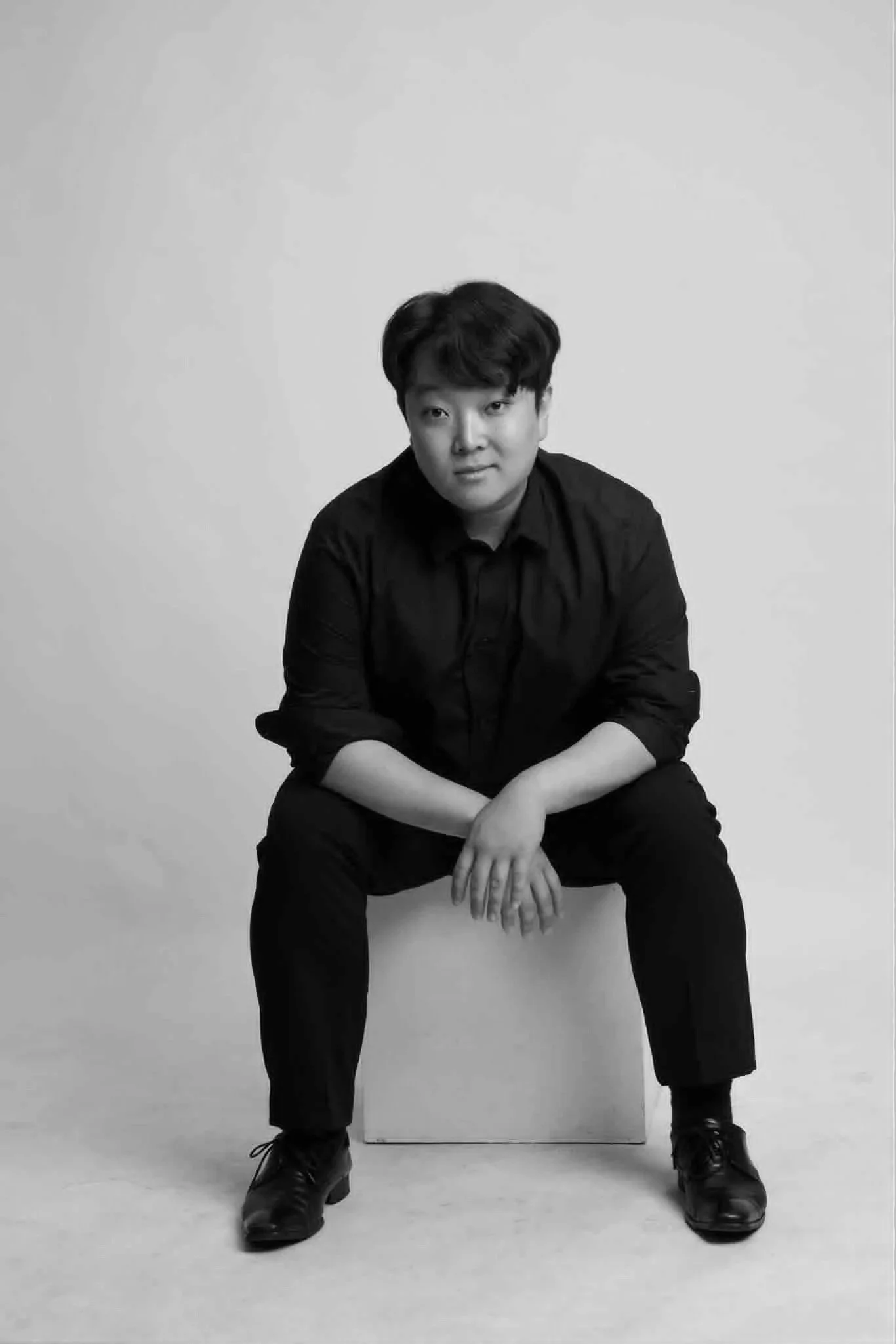 A black and white portrait of a young man sitting on a box against a plain background, wearing a dark shirt and pants, with his hands resting on his knees.