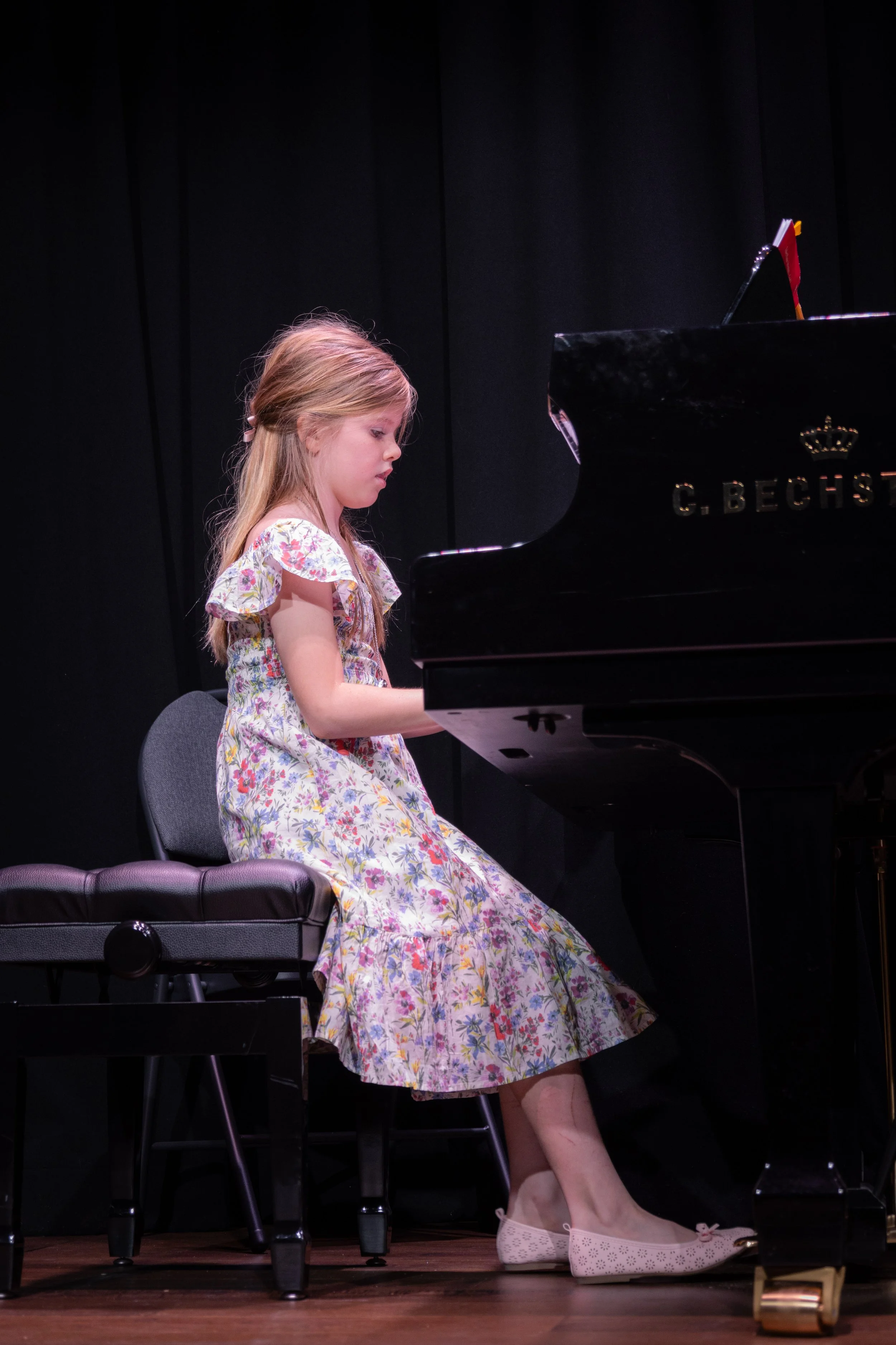 A young girl in a floral dress playing a grand piano on a stage.