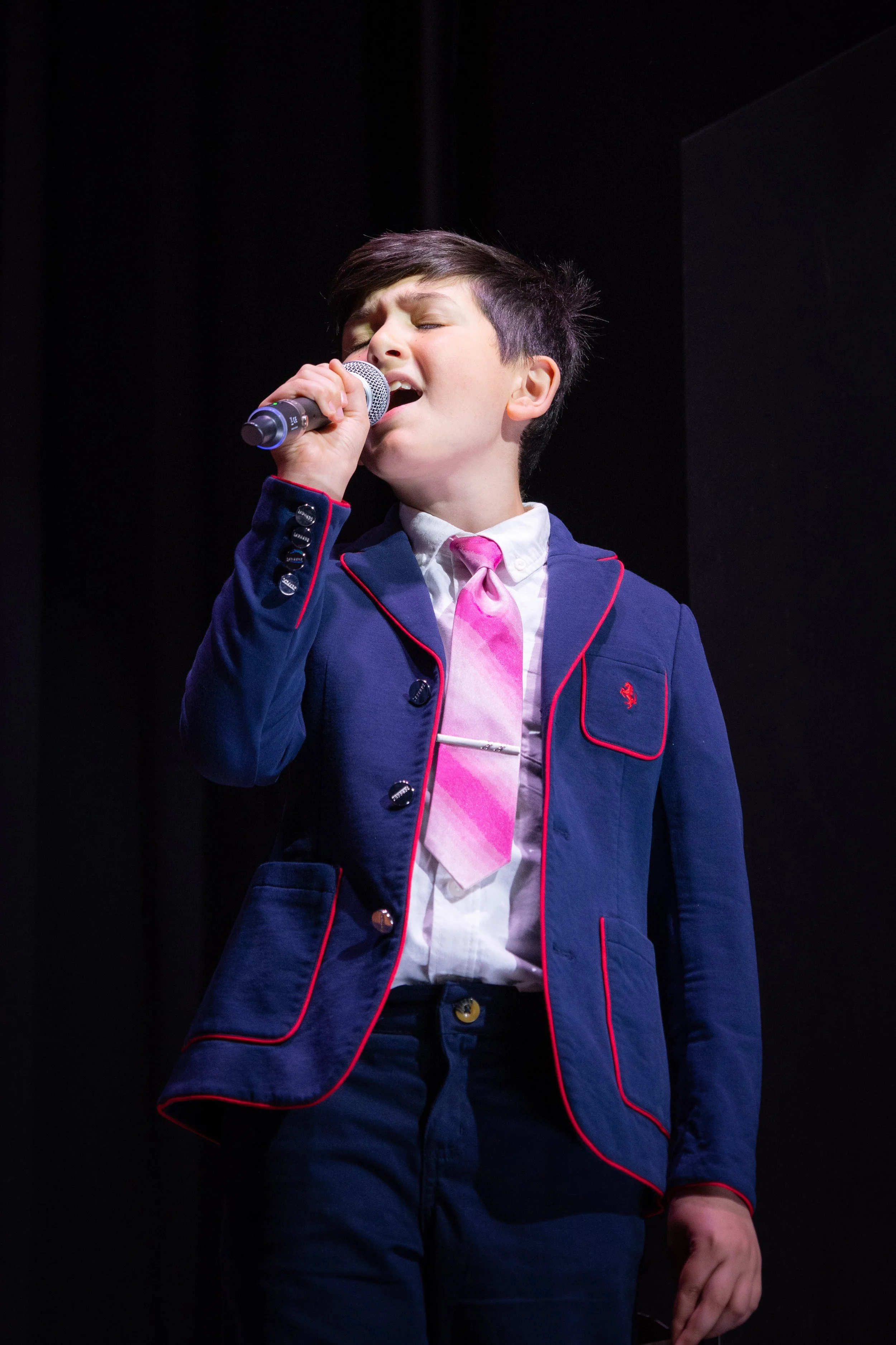 A young boy in a navy blazer with red piping, a white shirt, and a pink tie singing into a microphone on stage.