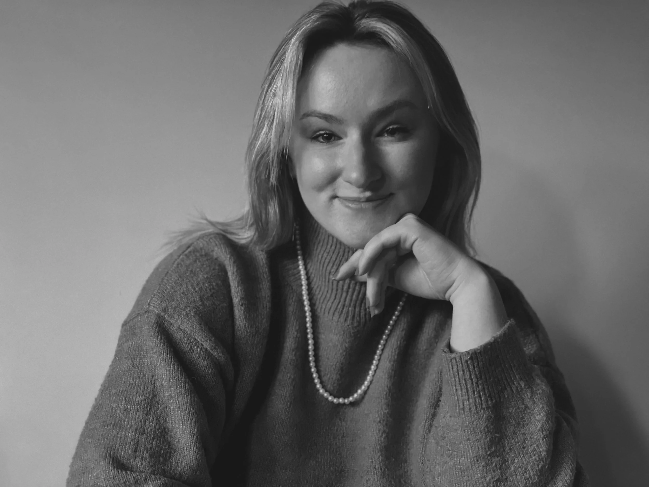 Black-and-white portrait of a woman with shoulder-length hair, smiling softly, wearing a sweater and a pearl necklace, resting her chin on her hand.