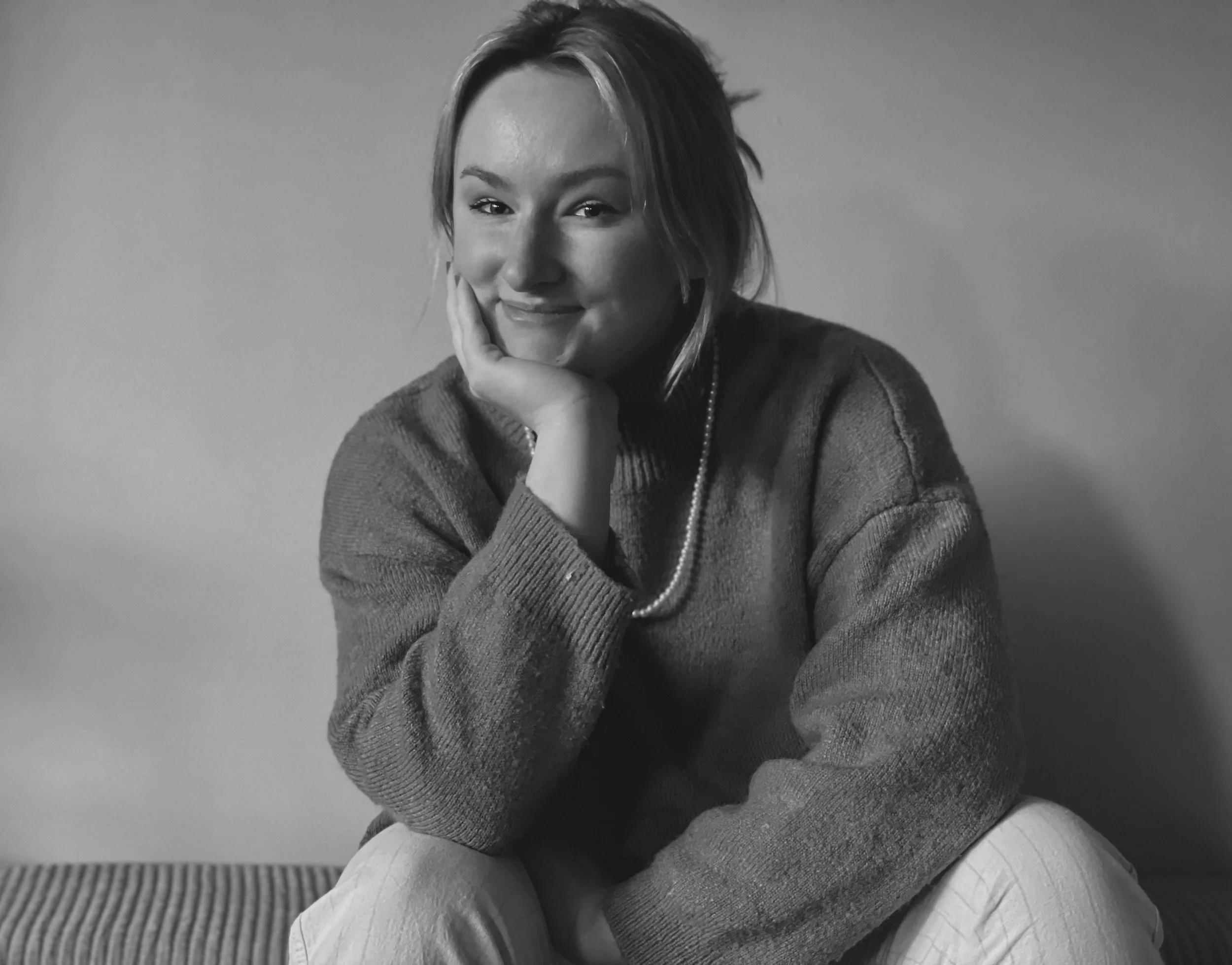 Black and white photo of a smiling woman sitting on a striped cushion, resting her chin on her hand, wearing a sweater and a pearl necklace.