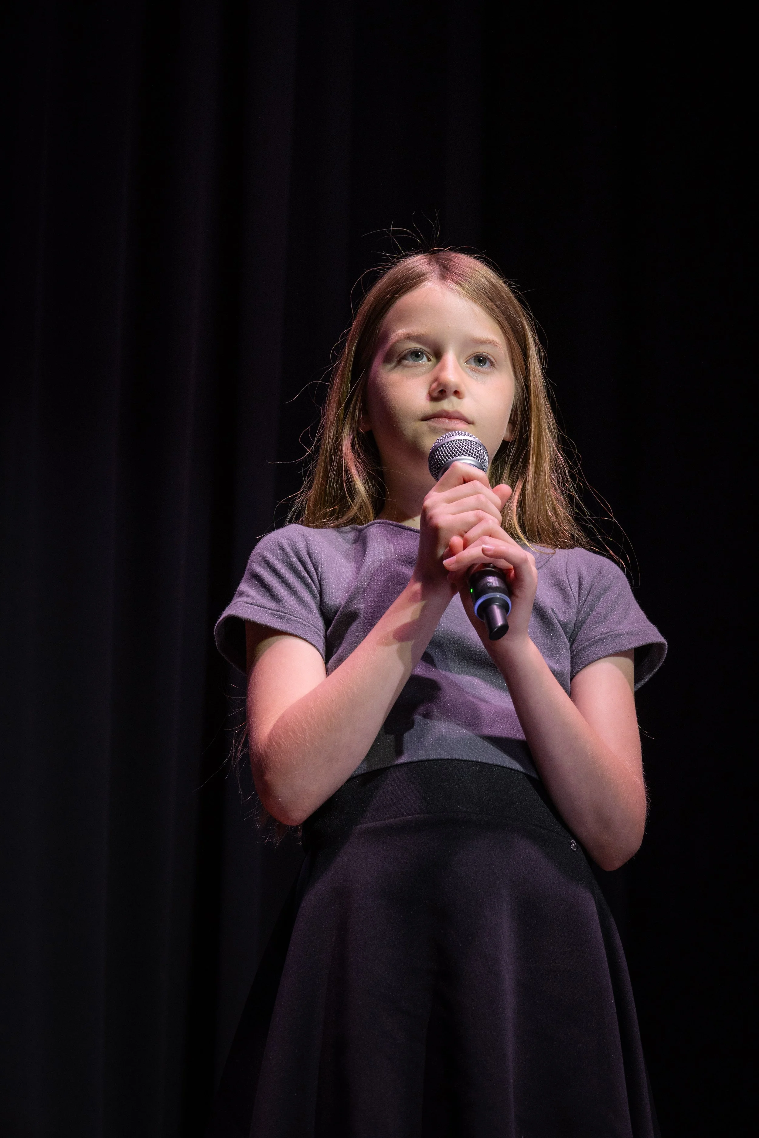 A young girl with long hair holding a microphone on stage against a black curtain background.