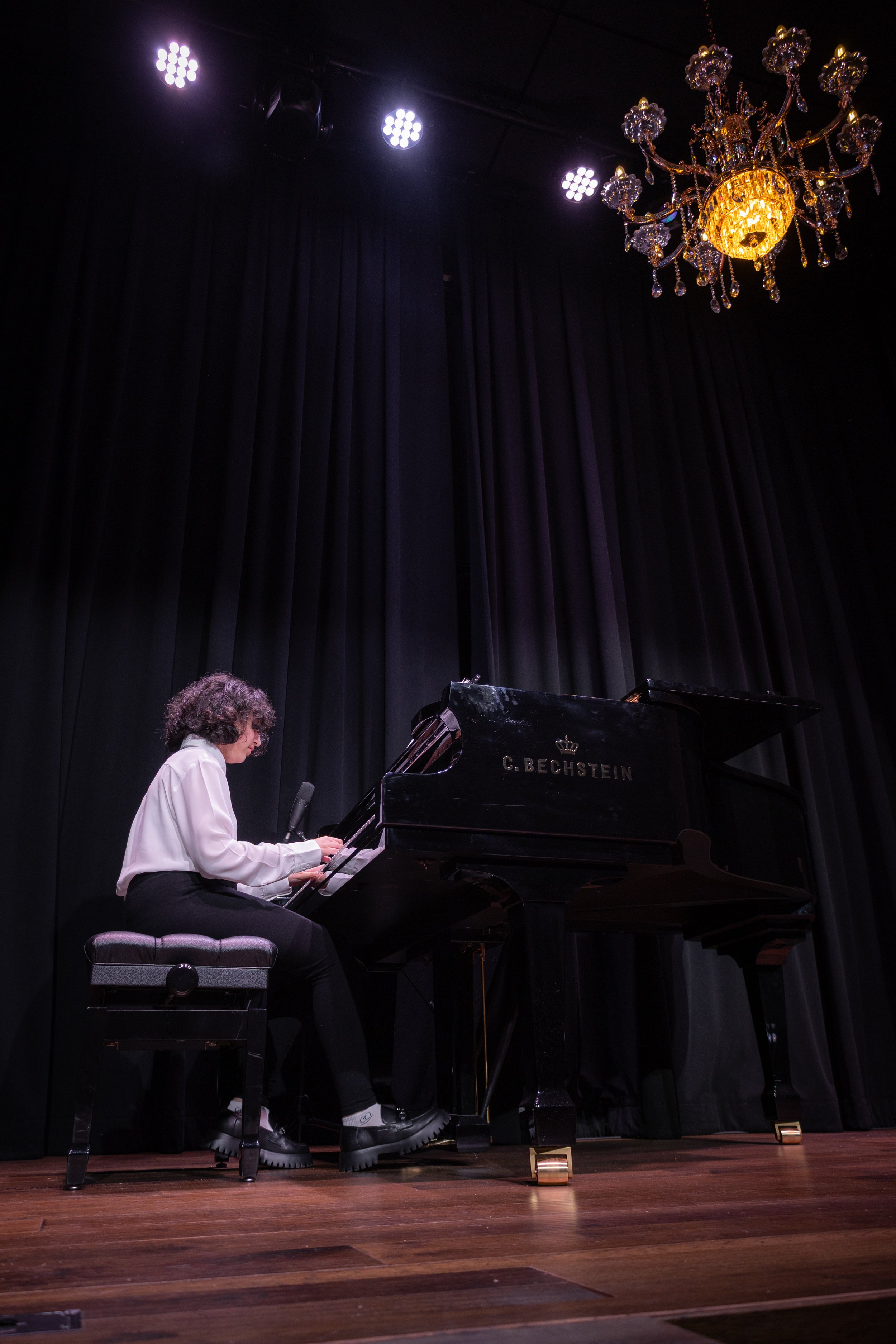 Person playing black grand piano on stage with black curtains, chandelier overhead, and stage lights.