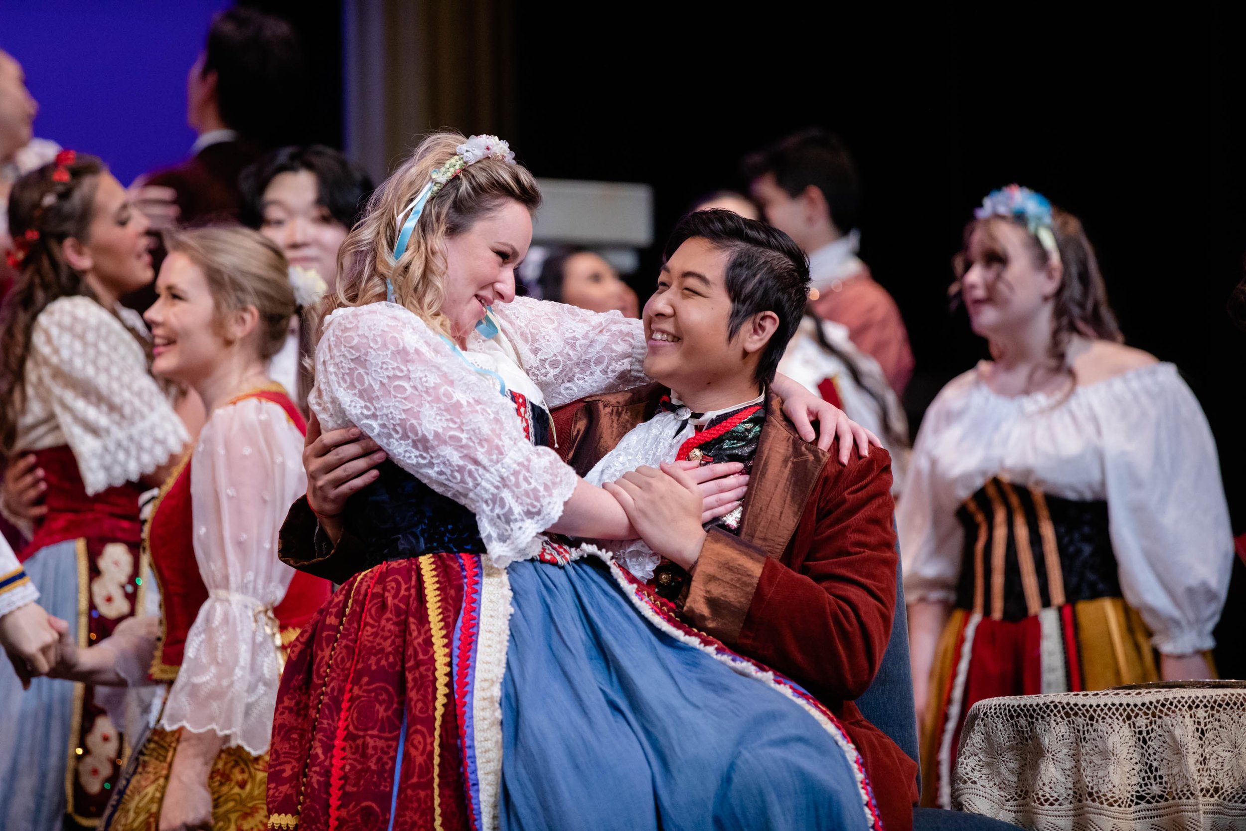 People dressed in traditional Scandinavian folk costumes performing a dance, with a woman sitting and a man holding her, smiling, surrounded by others in similar costumes.