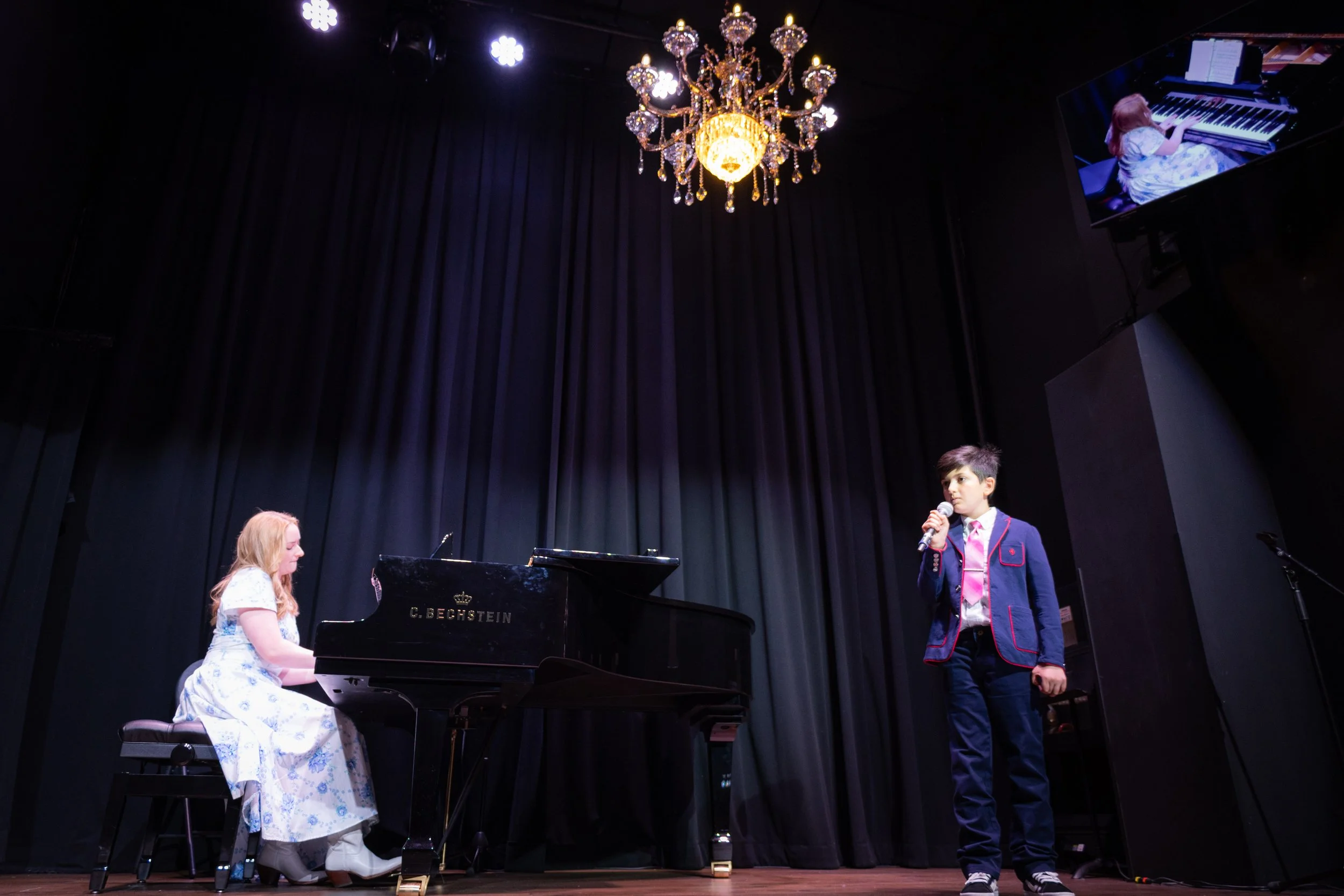 A young girl plays the piano on stage while a young boy stands holding a microphone, performing in a concert with black curtains and a decorative chandelier overhead.