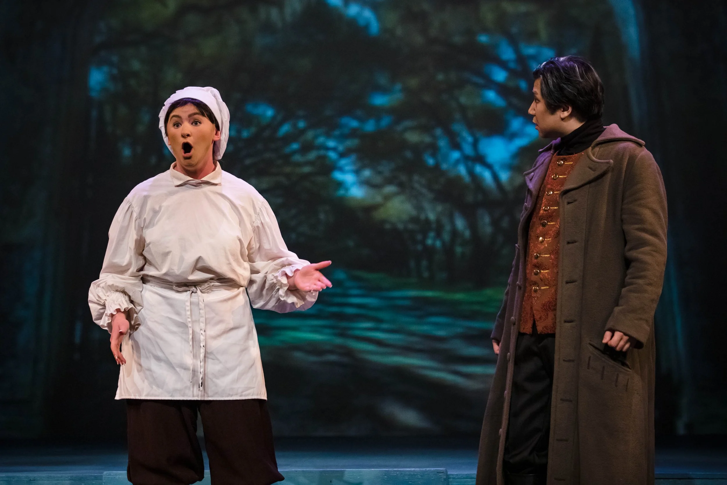 Two actors on stage, one dressed as a chef with a surprised expression, the other in a brown coat, set against a backdrop of a forest scene.