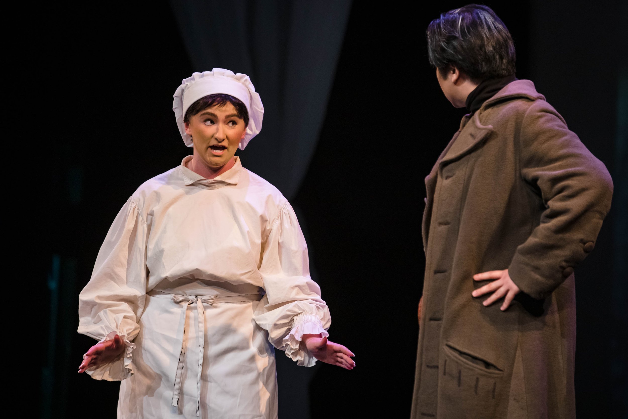 Two actors performing on stage, one dressed as a chef and the other in a brown coat, engaged in a theatrical scene.