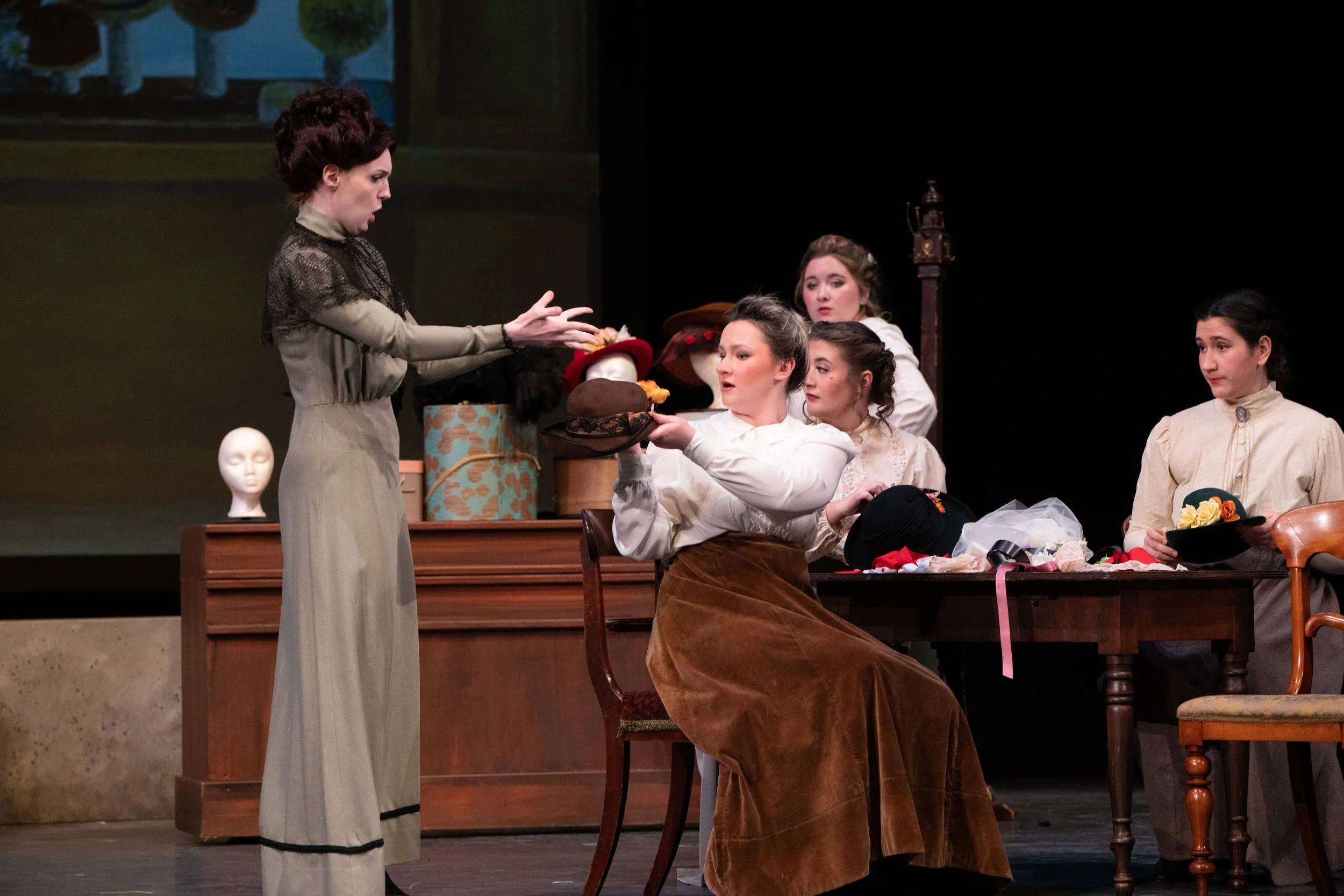 Six women in vintage clothing on stage, with hats and props, engaged in a theatrical performance.