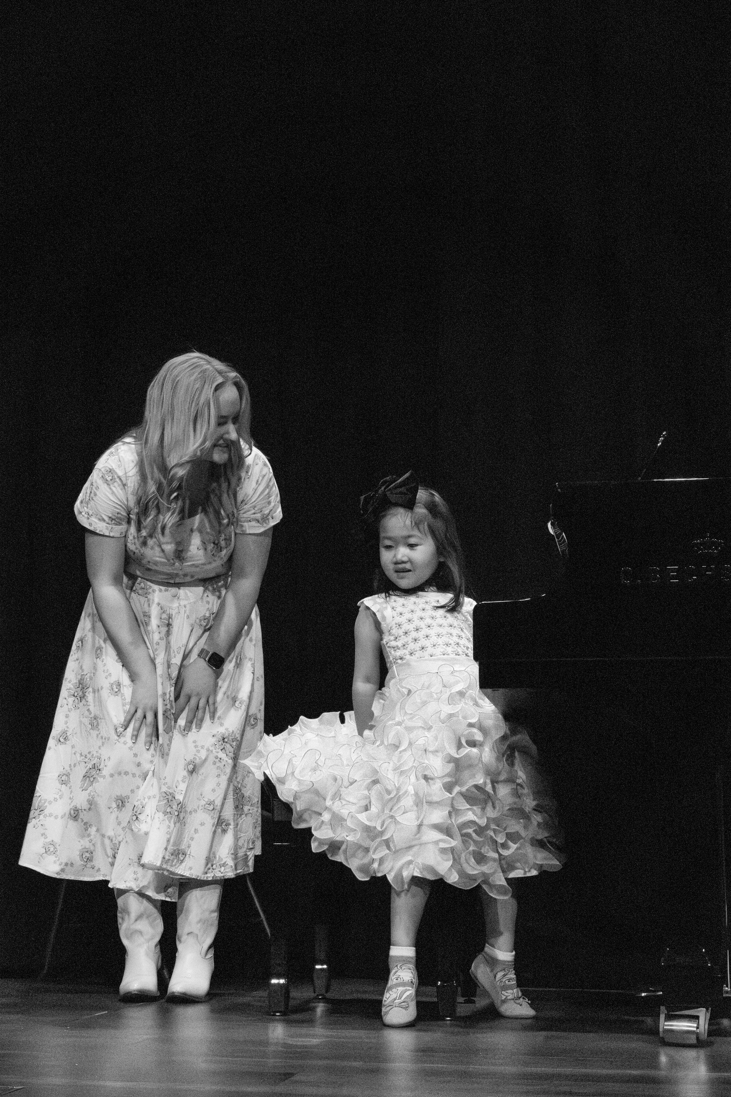 A woman and a young girl are standing next to a grand piano on a stage. The girl is wearing a fancy dress with ruffled skirt and the woman is wearing a floral dress and boots, and they are both smiling.