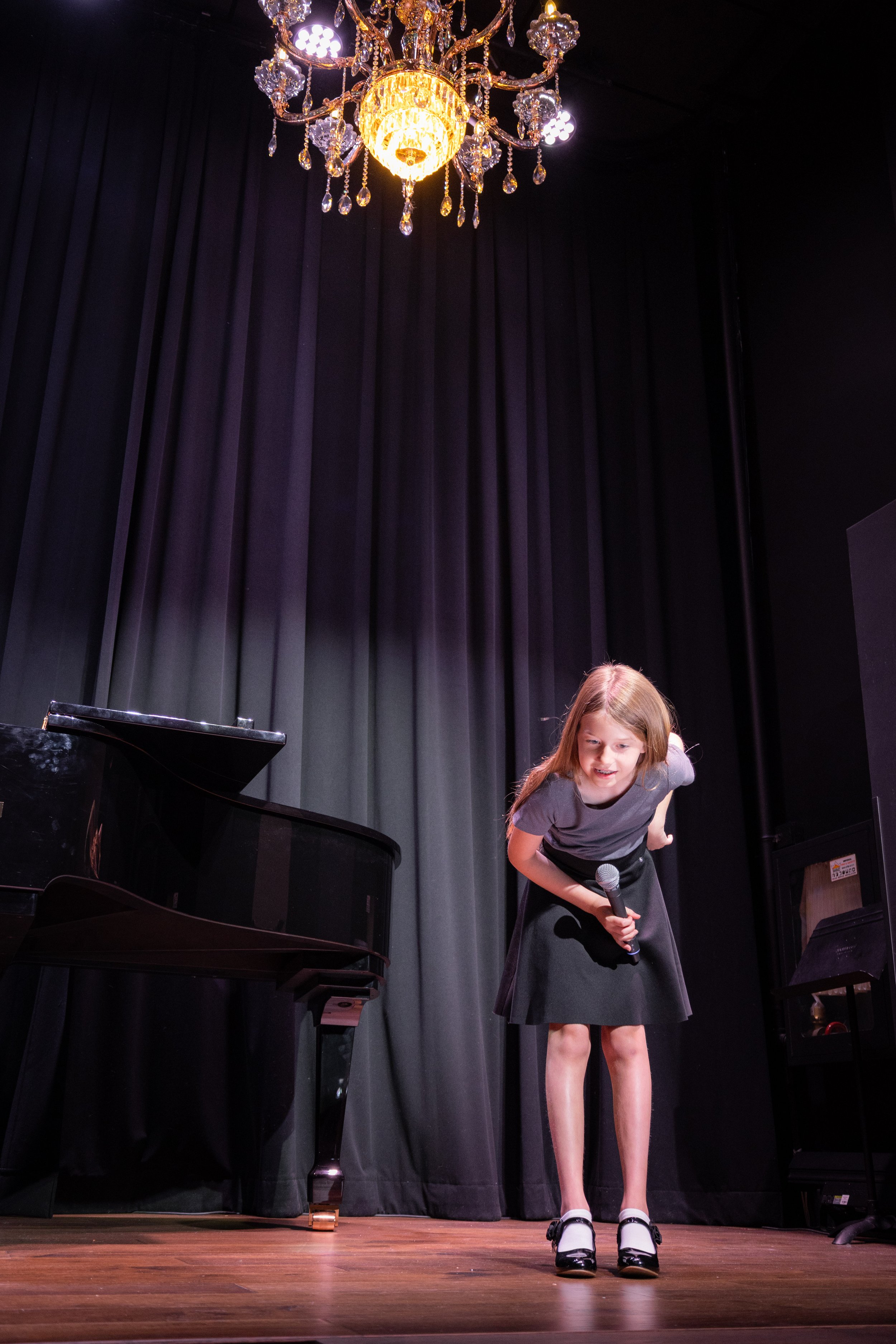 A young girl on stage holding a microphone, bowing while smiling, next to a black grand piano with black curtains in the background and a chandelier hanging from above.