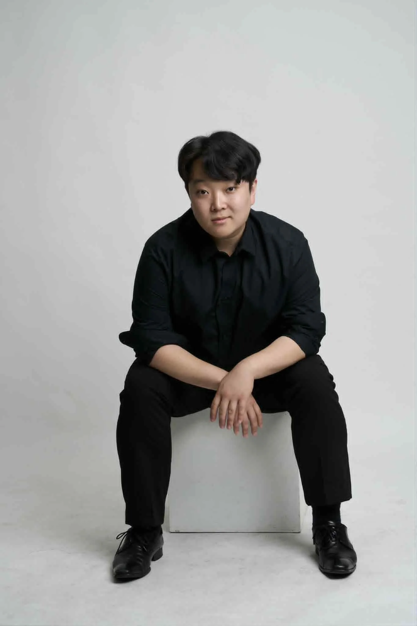A young man with dark hair, wearing a black shirt and black pants, sitting on a white block against a plain light background.