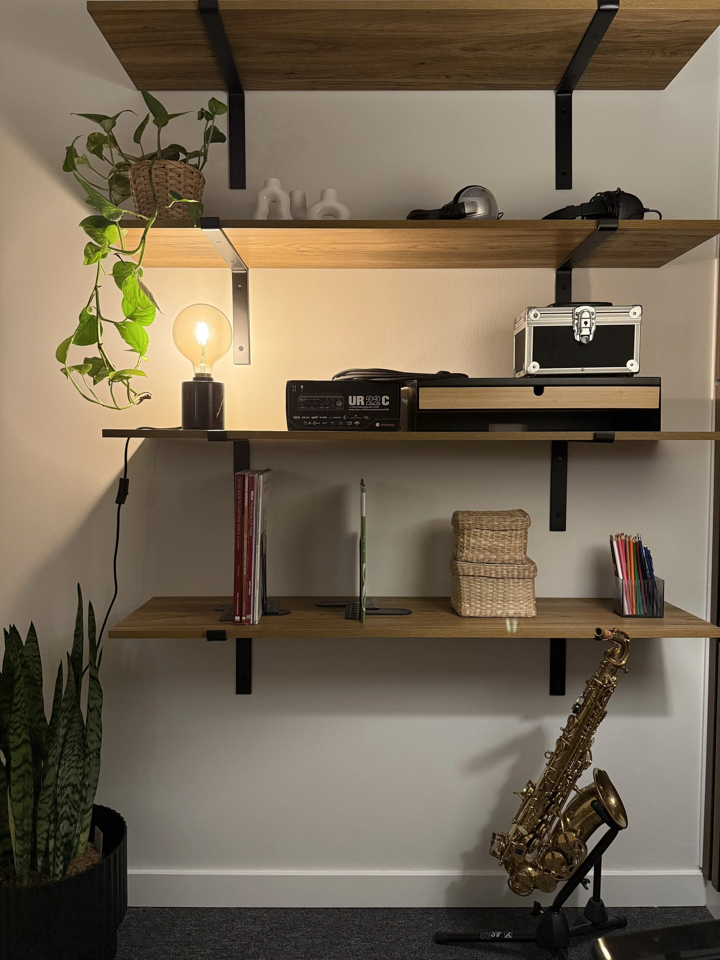 Wall-mounted shelves holding potted plant, decorative vases, headphones, a lightbulb lamp, audio equipment, books, baskets, colored pencils, and a saxophone on a stand.