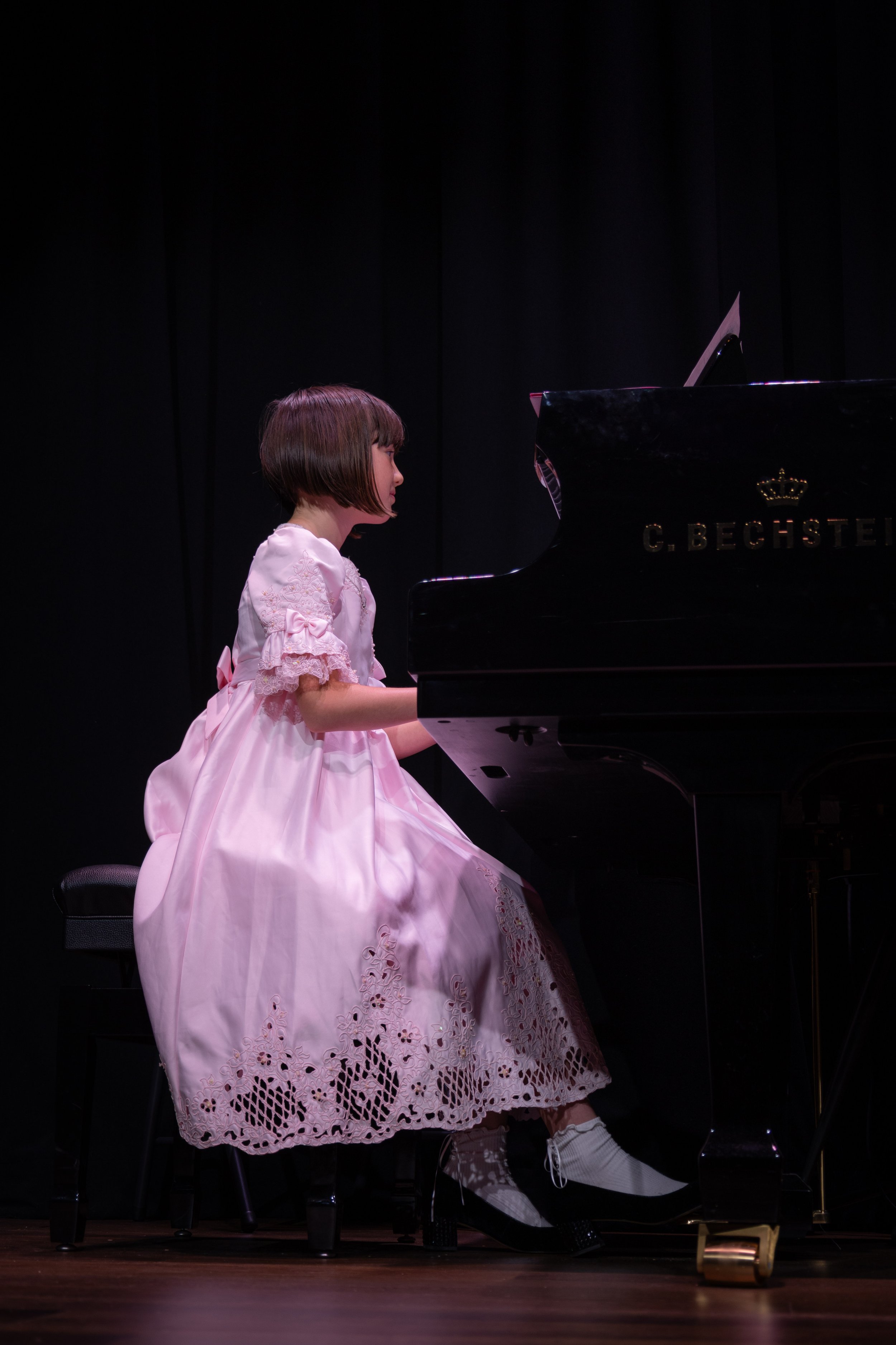 A young girl in a pink dress playing a black grand piano on a dark stage.