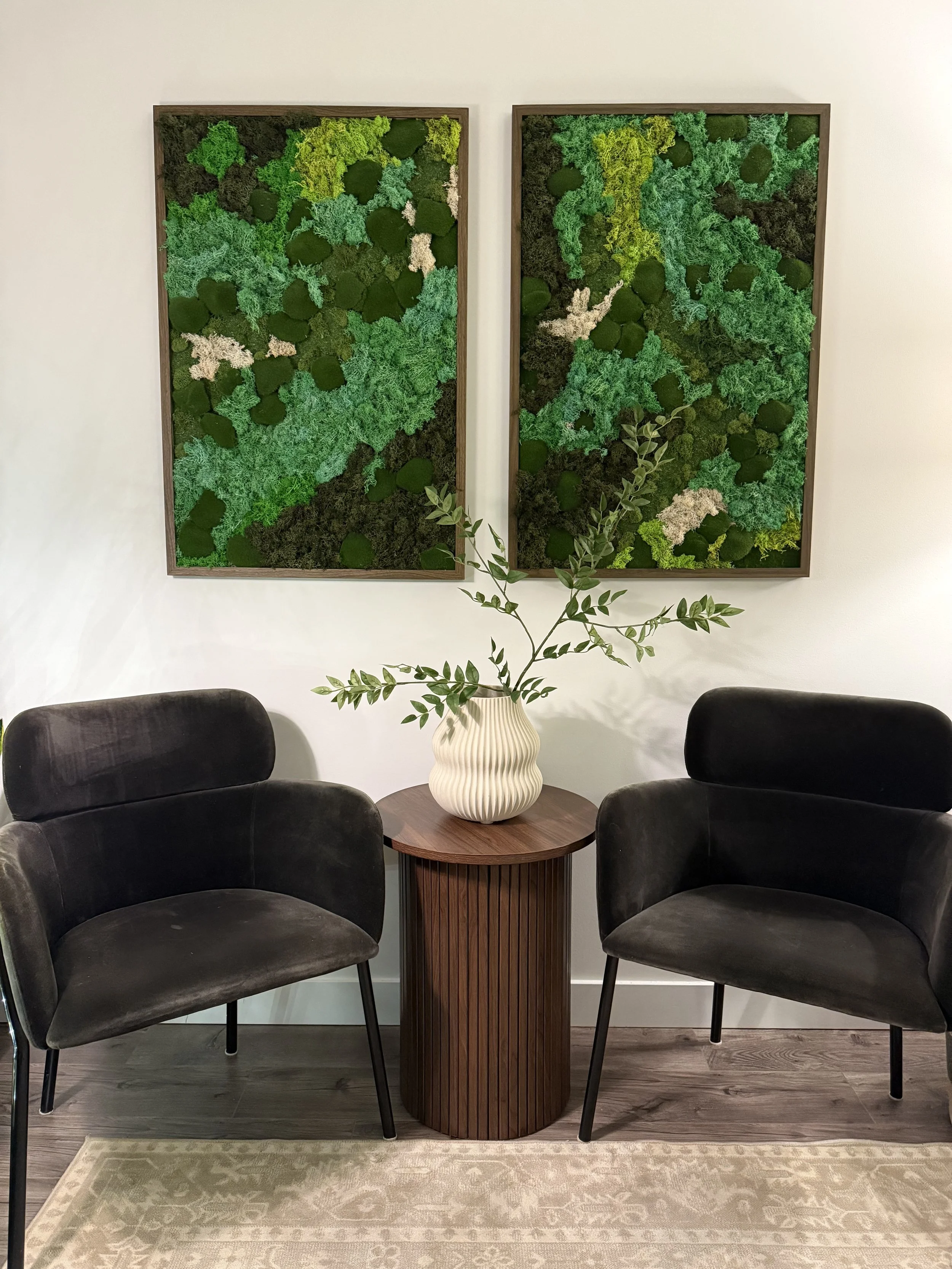 Two black armchairs on each side of a round wooden table with a white ribbed vase containing green leaves, with two green moss wall art pieces hanging on the white wall behind, and a beige rug on the dark wood floor.