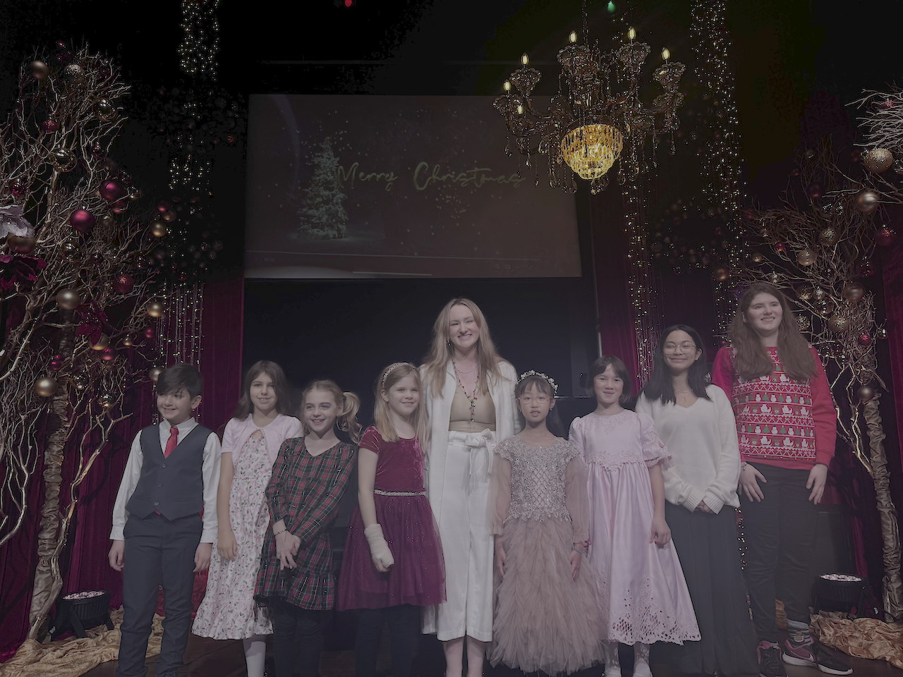 Group of children and an adult woman on a decorated stage for a Christmas celebration, with festive trees, lights, and Christmas decorations.