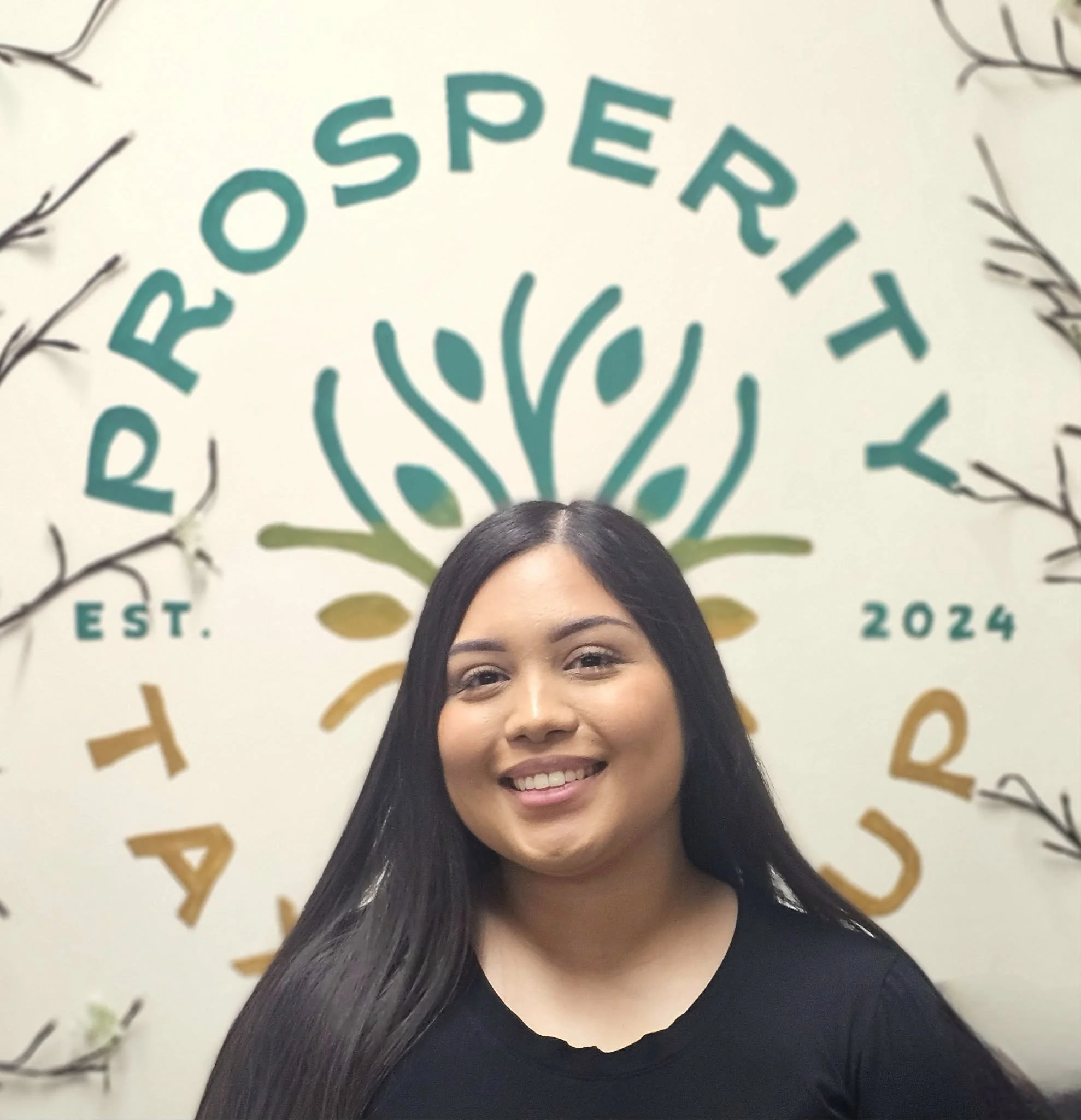 A woman with long black hair, wearing a black top and a silver cross necklace, stands in front of a wall with a sign that reads 'ROOSEPRIETY' and 'EST. 2024', surrounded by decorative string lights and greenery.