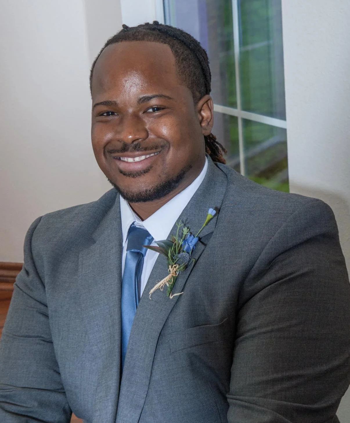 Man in gray suit with blue tie and boutonniere, smiling indoors near window.