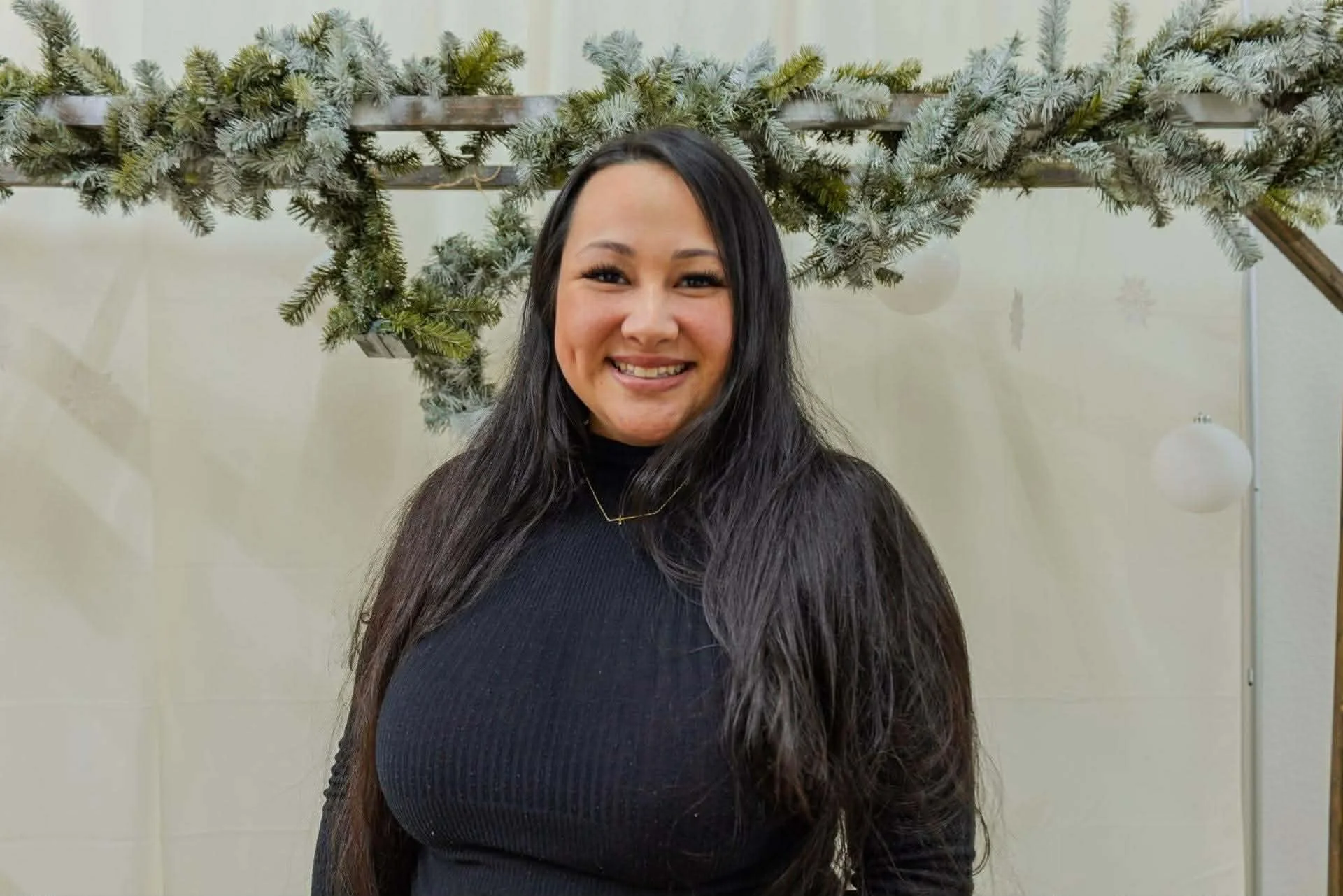 A woman with long black hair smiling at the camera, wearing a black top, standing in front of holiday decorations including frosted pine branches and hanging ornaments.