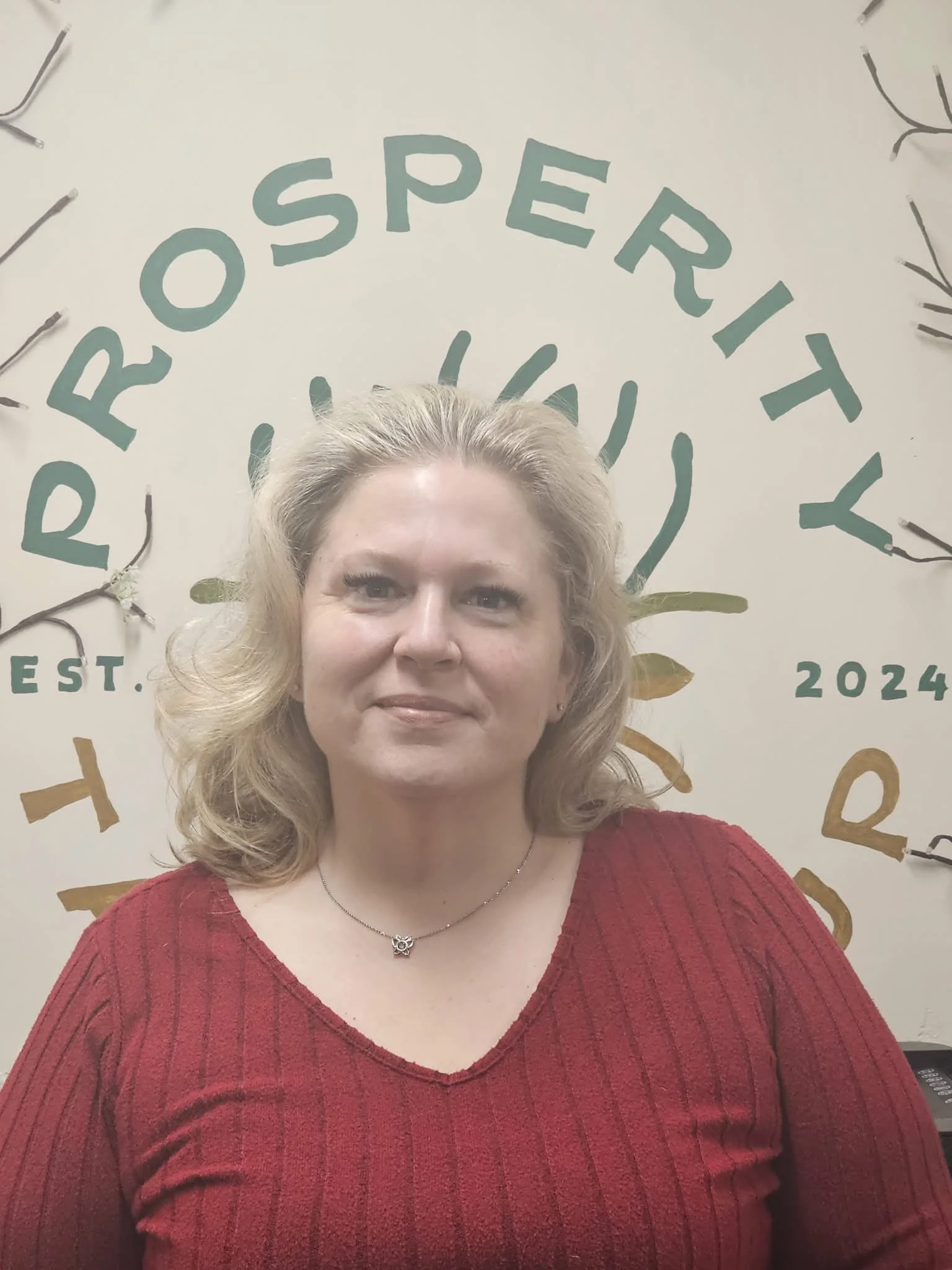 A woman with shoulder-length blonde hair wearing a red sweater and a necklace standing in front of a wall with the words "IROROSPEERITTY" and the year "2024" written on it, along with decorative branches.