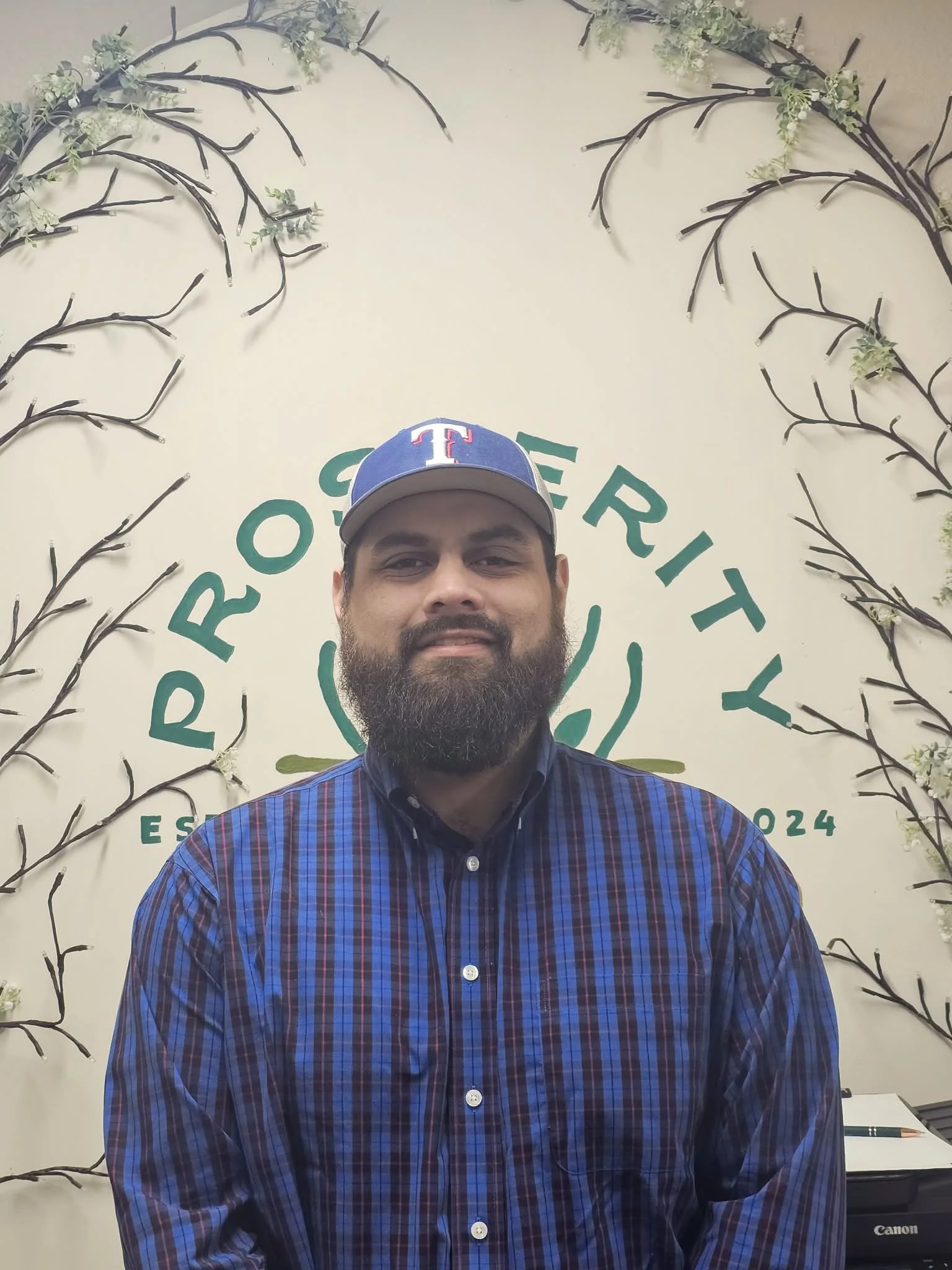 A man with a beard wearing a Texas Rangers cap and a blue checkered shirt standing in front of a decorative background with branches and a logo for Rooster Ritz.