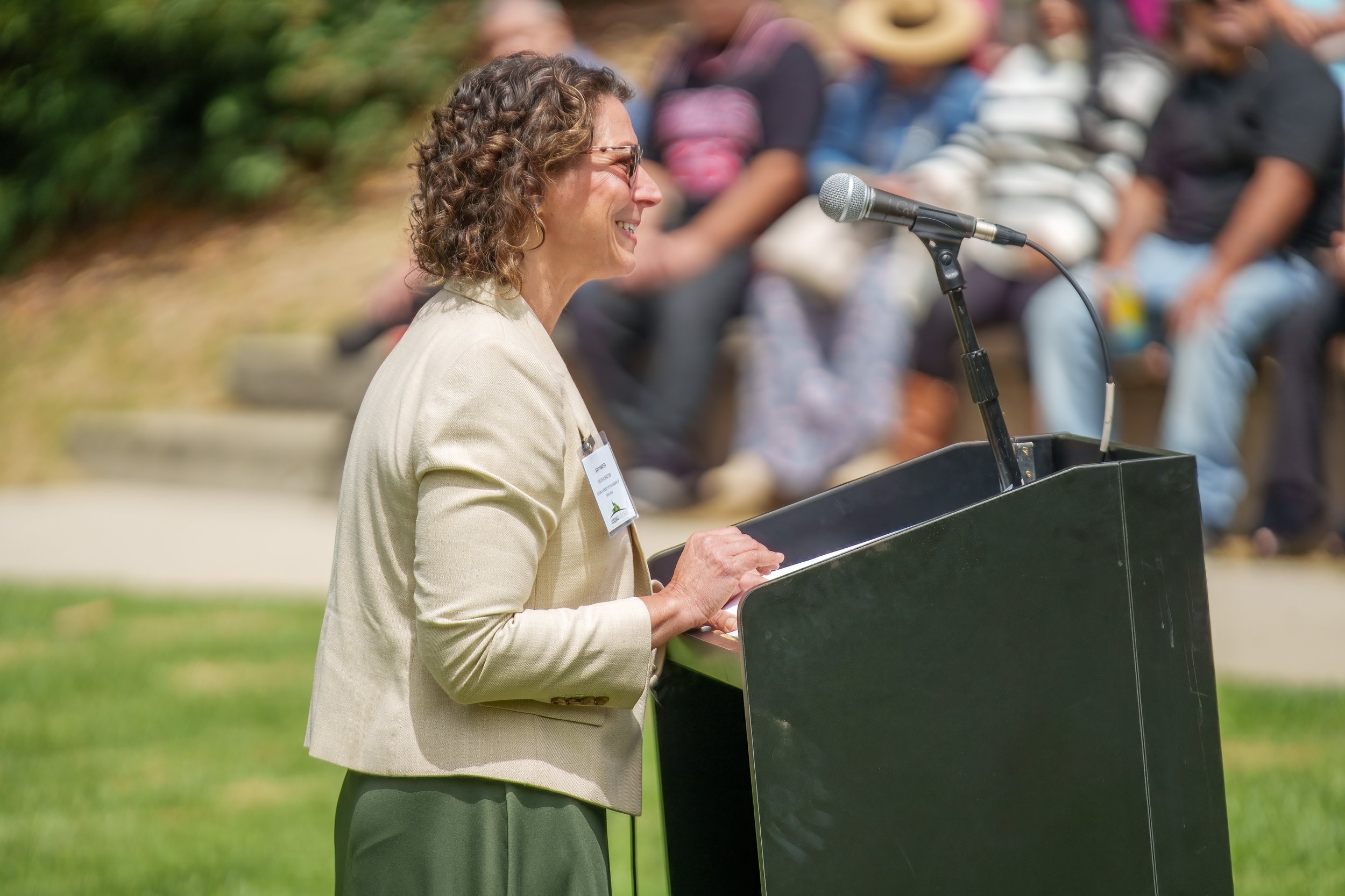A woman with curly hair and glasses speaking at a podium outdoors during a public event, with a microphone and an audience sitting behind her on a grassy area.