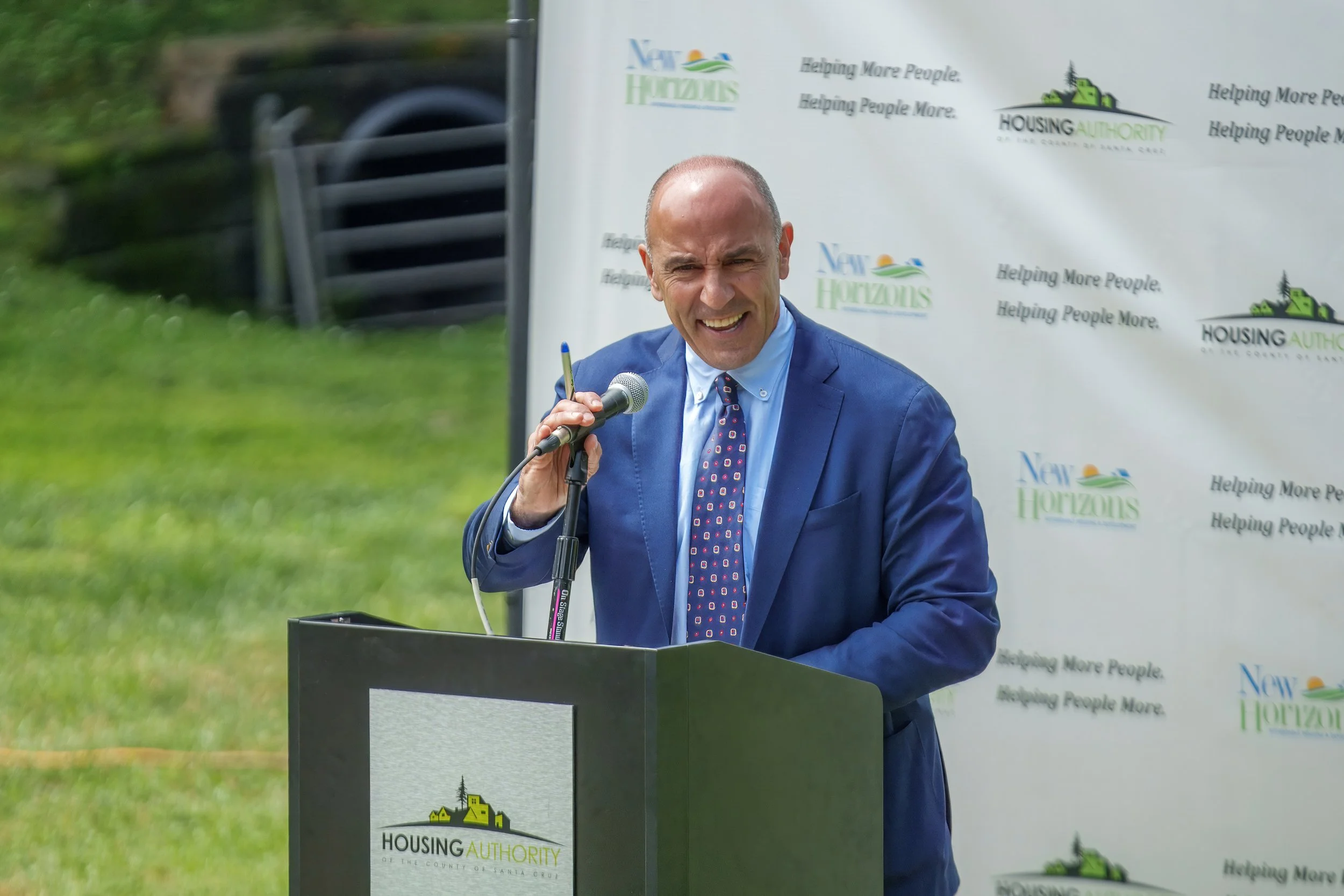 A man in a blue suit and tie speaking at a podium outdoors during an event. The background features a banner with the logos and slogans of the Housing Authority and New Horizons, and the grass is visible behind him.