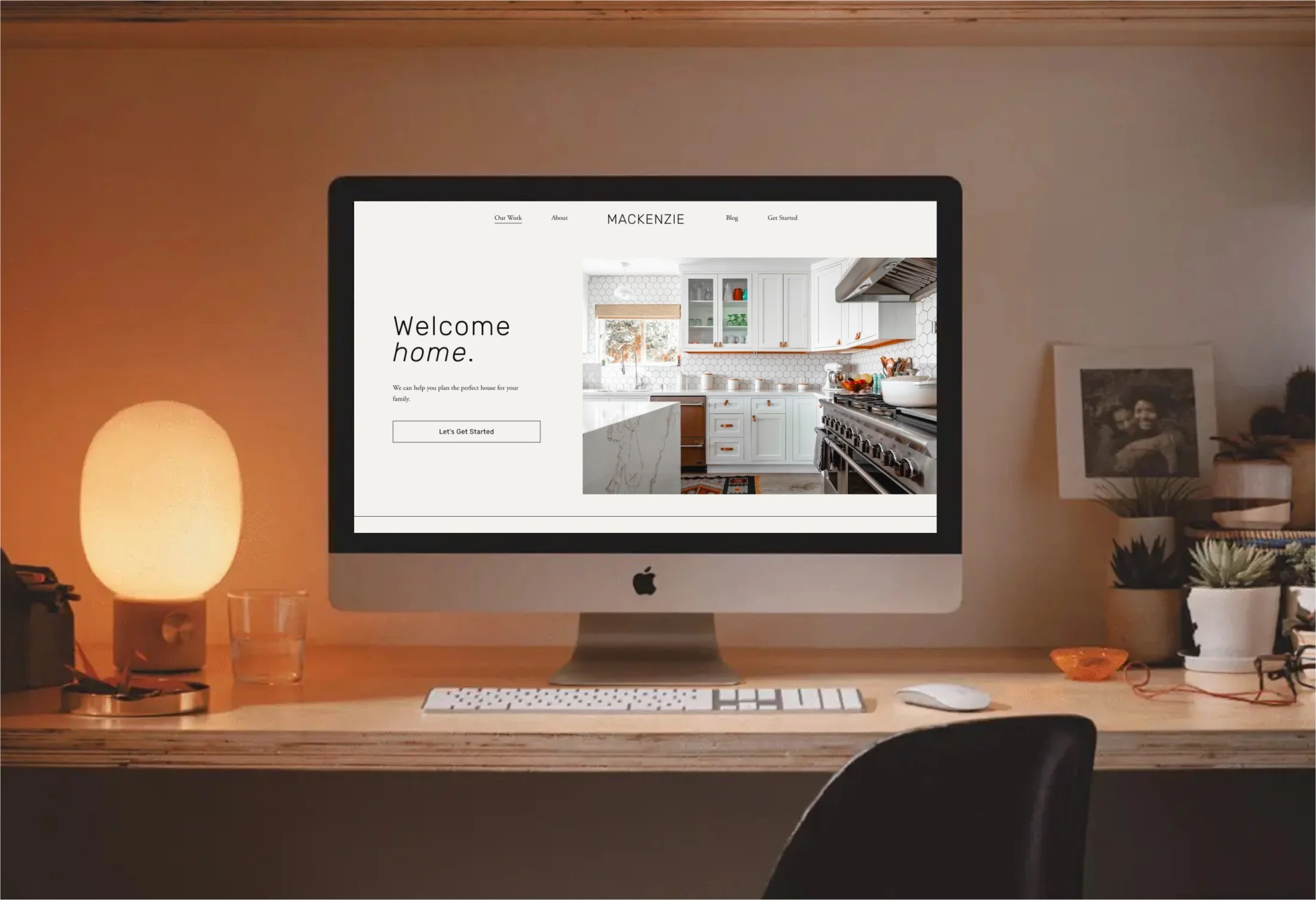 Computer monitor displaying a website for home design or real estate, with a kitchen image on the screen, on a wooden desk with a lamp, glass of water, and potted plants nearby.