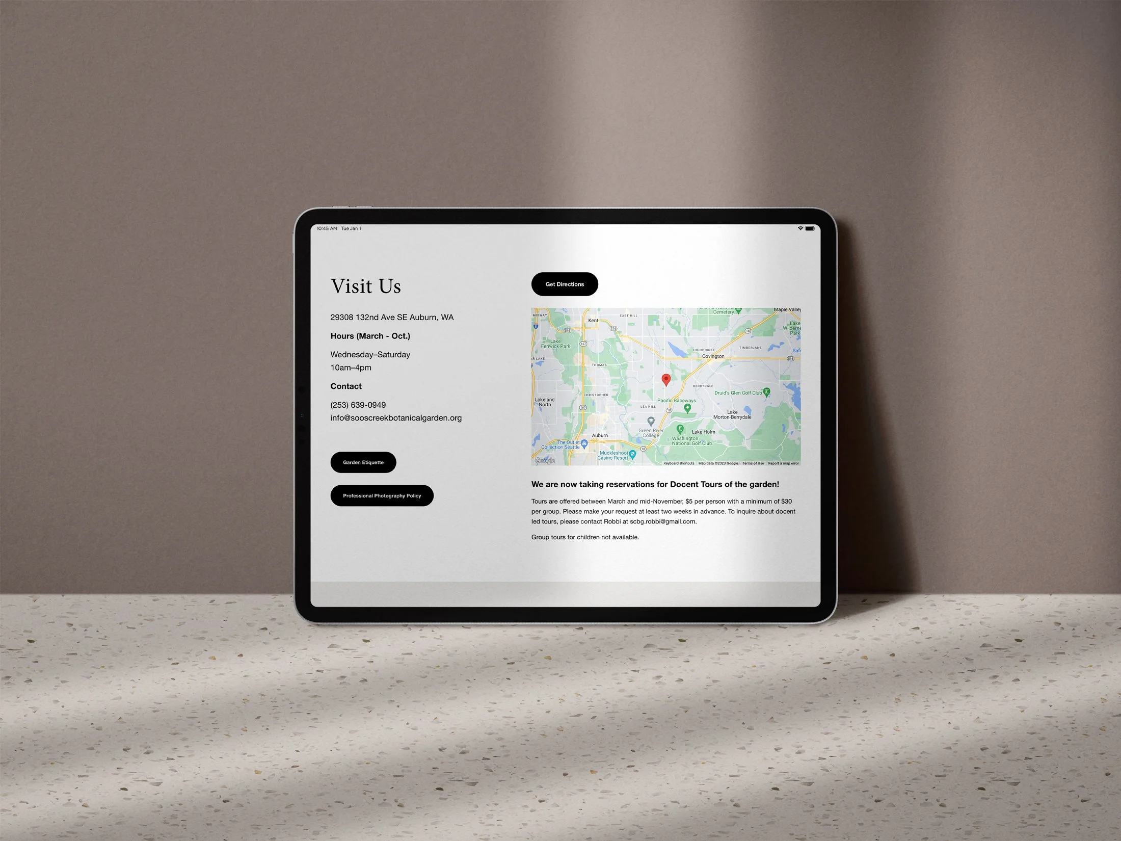Tablet displaying a map and contact information for a botanical garden, set on a speckled countertop against a plain wall.