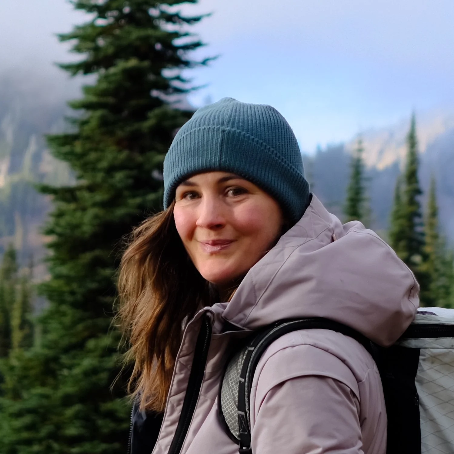 A woman with long brown hair wearing a blue knit cap and a beige outdoor jacket, smiling while hiking in a forested mountainous area with tall trees and mountains in the background.
