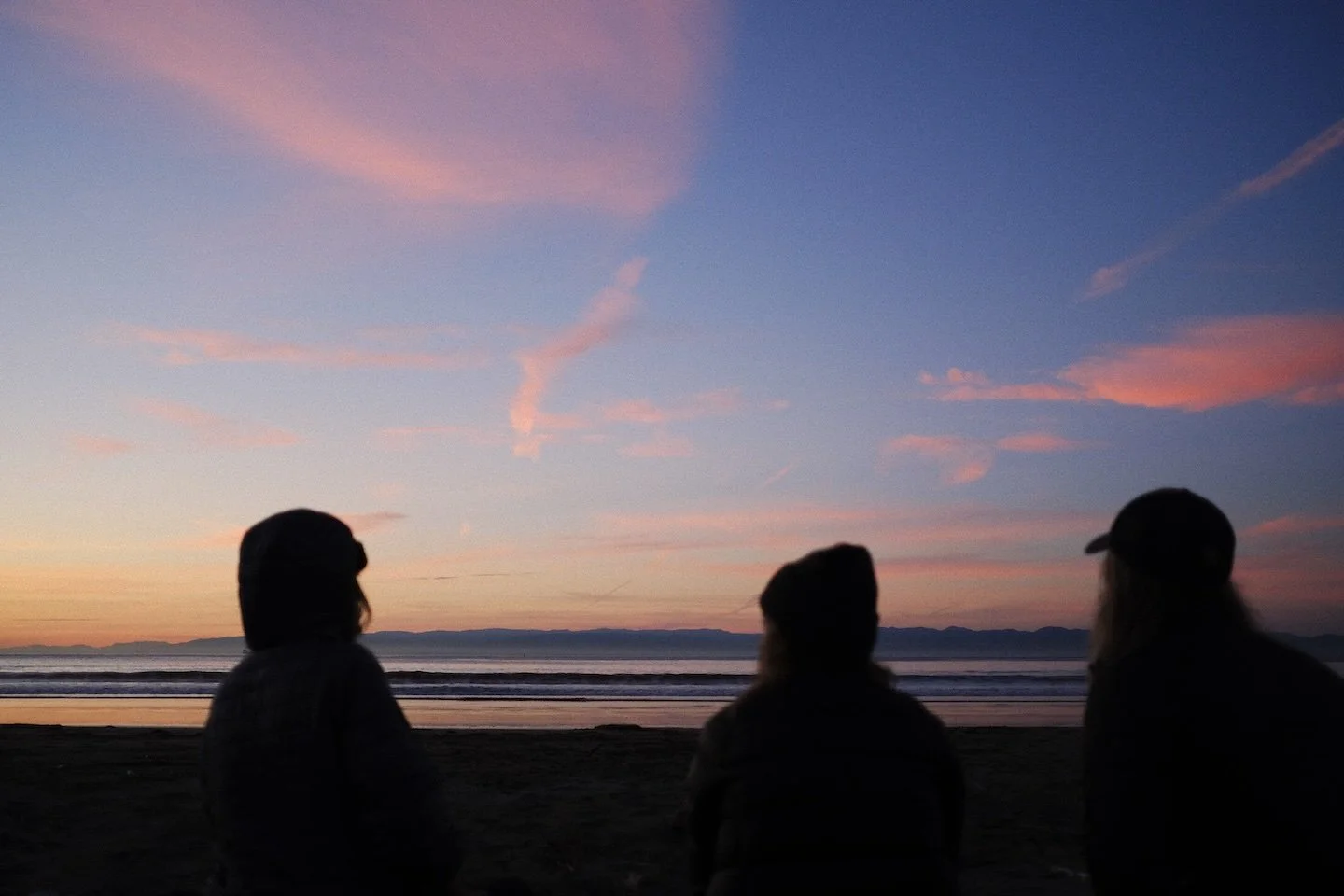 Three silhouettes of people sitting on a beach at sunset, with a colorful sky and distant mountains over the ocean.