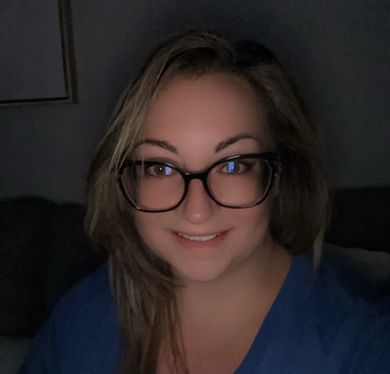 A smiling woman with brown hair, glasses, and wearing a blue top, sitting in a dark room.