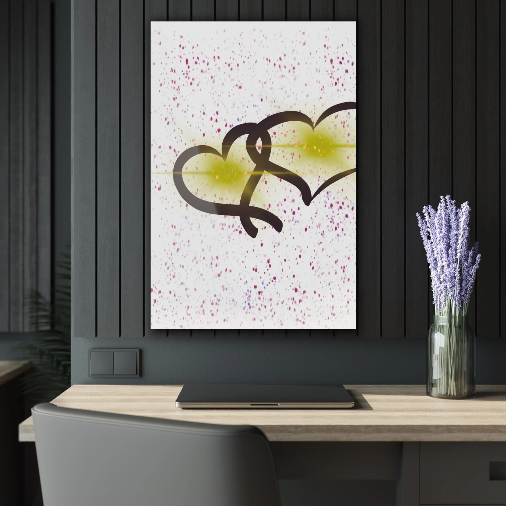 Wall art painting with two intertwined hearts, black outline, yellow accents, on a white background with pink and purple splatters, on a dark wood paneled wall above a wooden desk with a laptop and a glass vase of purple lavender flowers.
