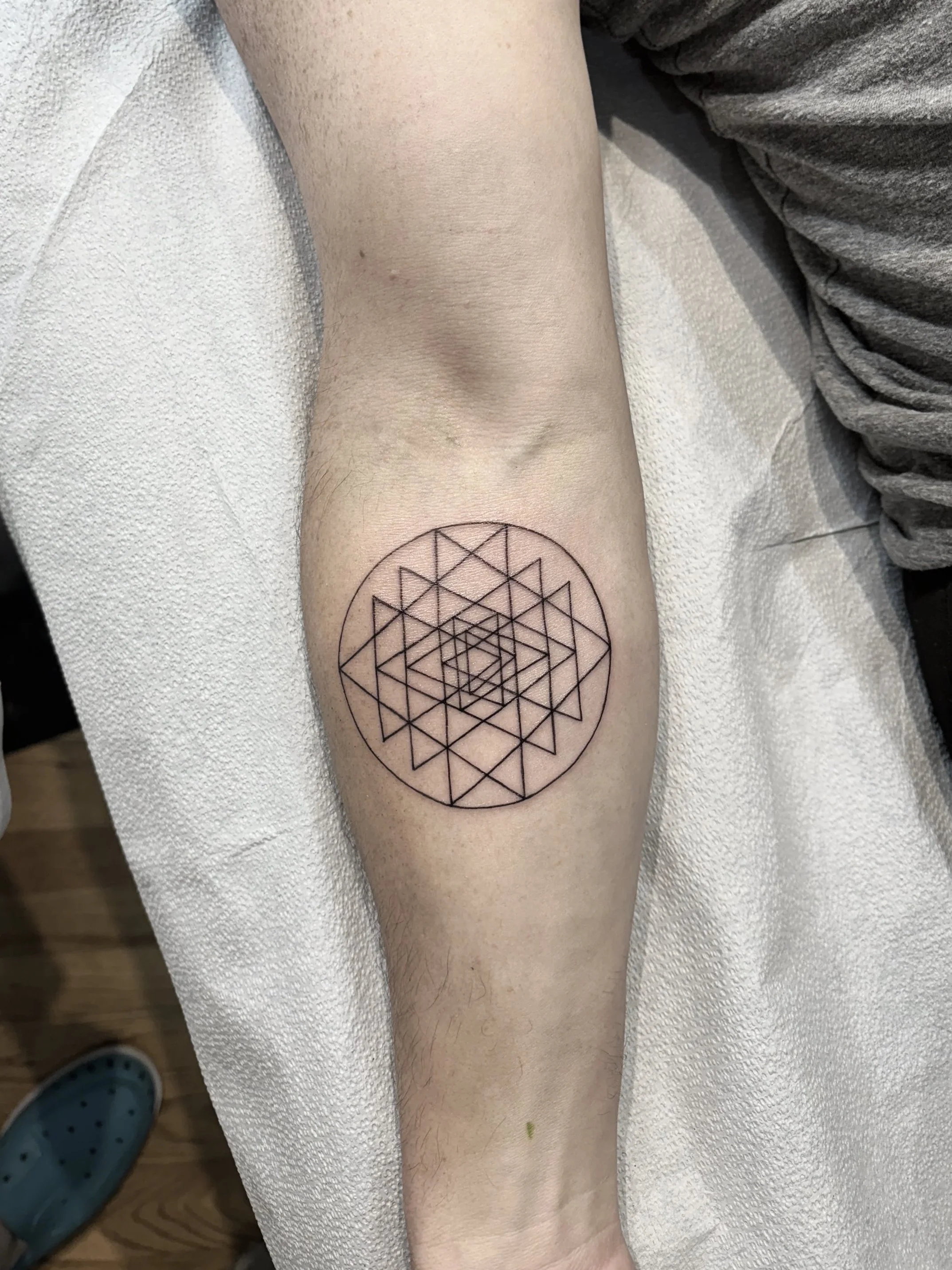 A person's forearm with a geometric tattoo design, resting on a white textured surface, with a gray shirt sleeve visible, and a shoe with holes seen partially at the bottom.