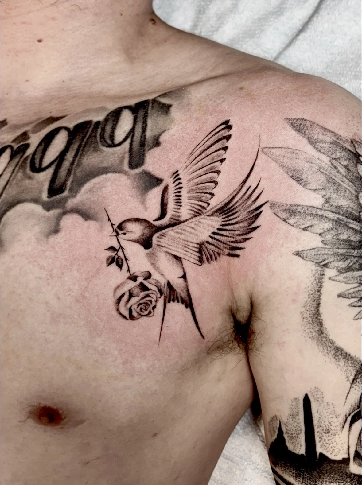 Tattoo on person's chest depicting a hummingbird with a rose in its beak, surrounded by feathers, clouds, and text.