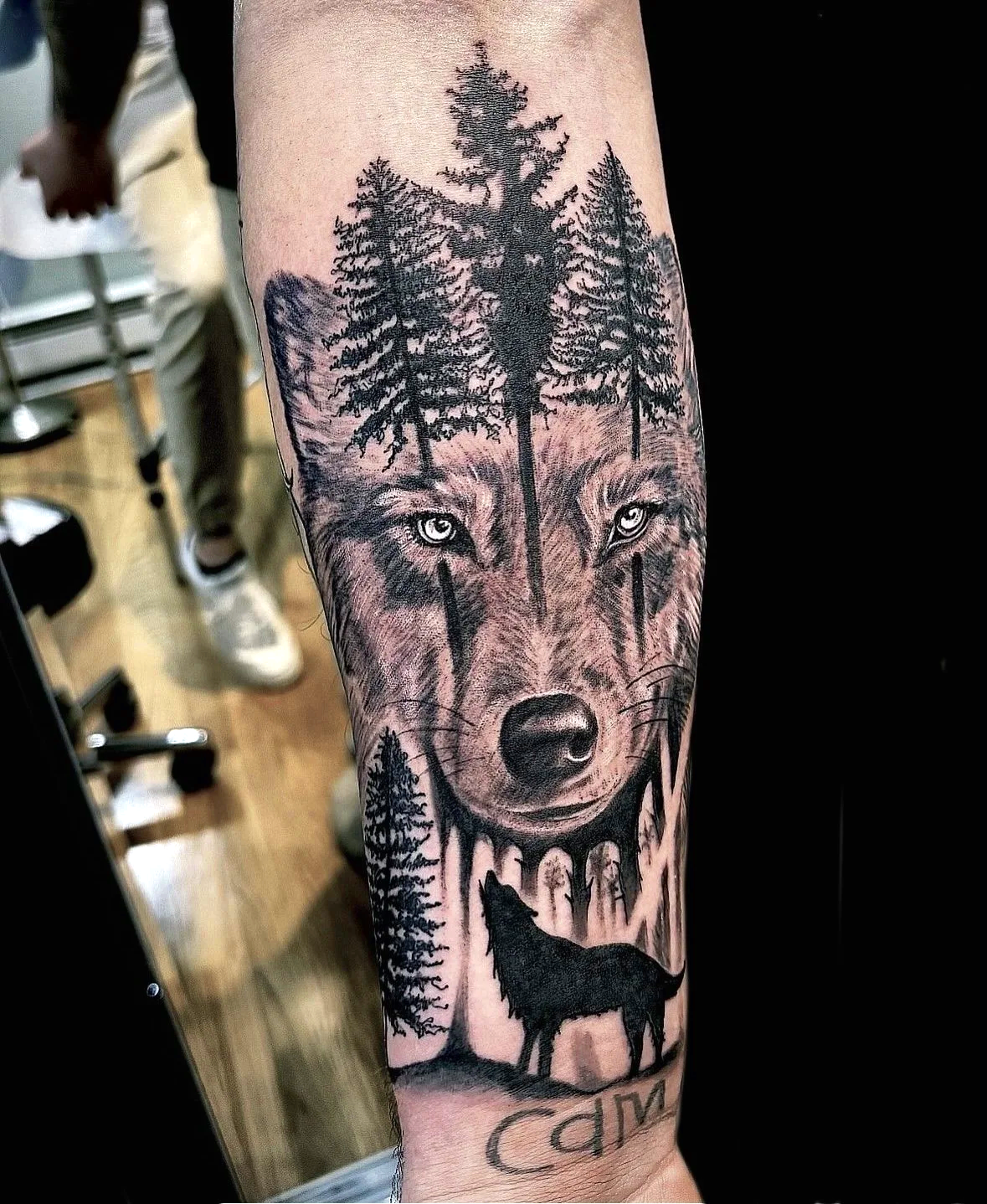 Tattoo of a wolf's face with piercing eyes, trees growing from its head, and a howling wolf silhouette at the bottom.