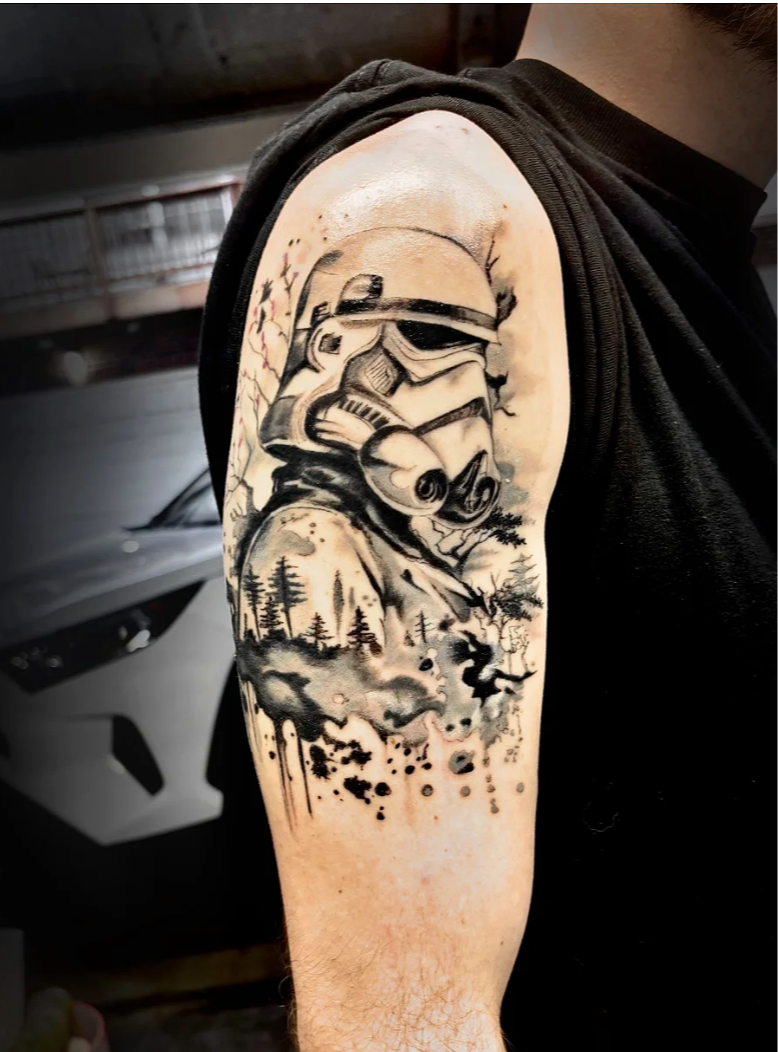 Black and gray tattoo of a Stormtrooper helmet from Star Wars emerging from a landscape with trees and smoke on the upper arm.