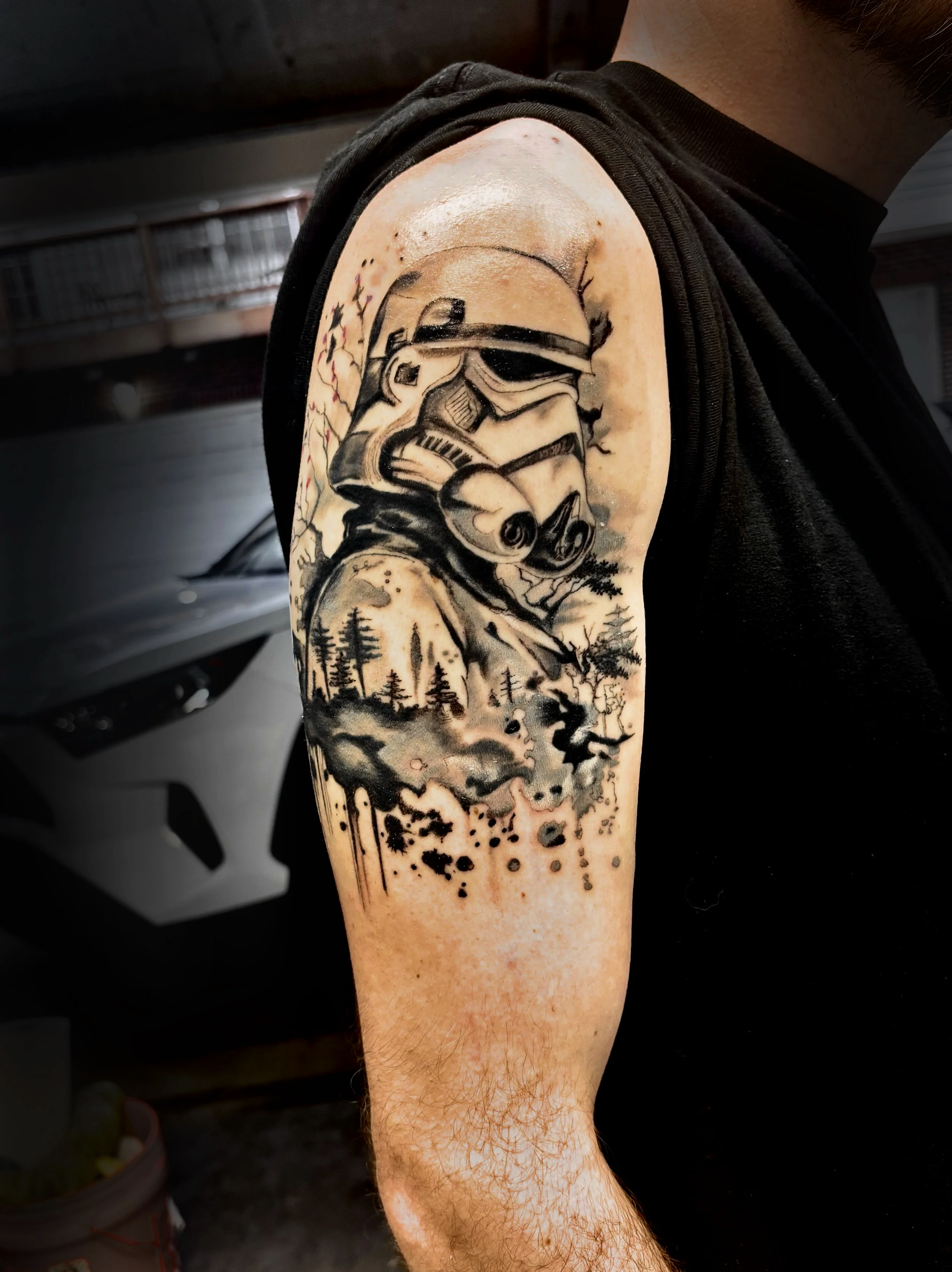 Black and gray tattoo of a stormtrooper helmet from Star Wars, with a background of trees and a cloudy sky, on the upper arm of a person with light skin and arm hair.